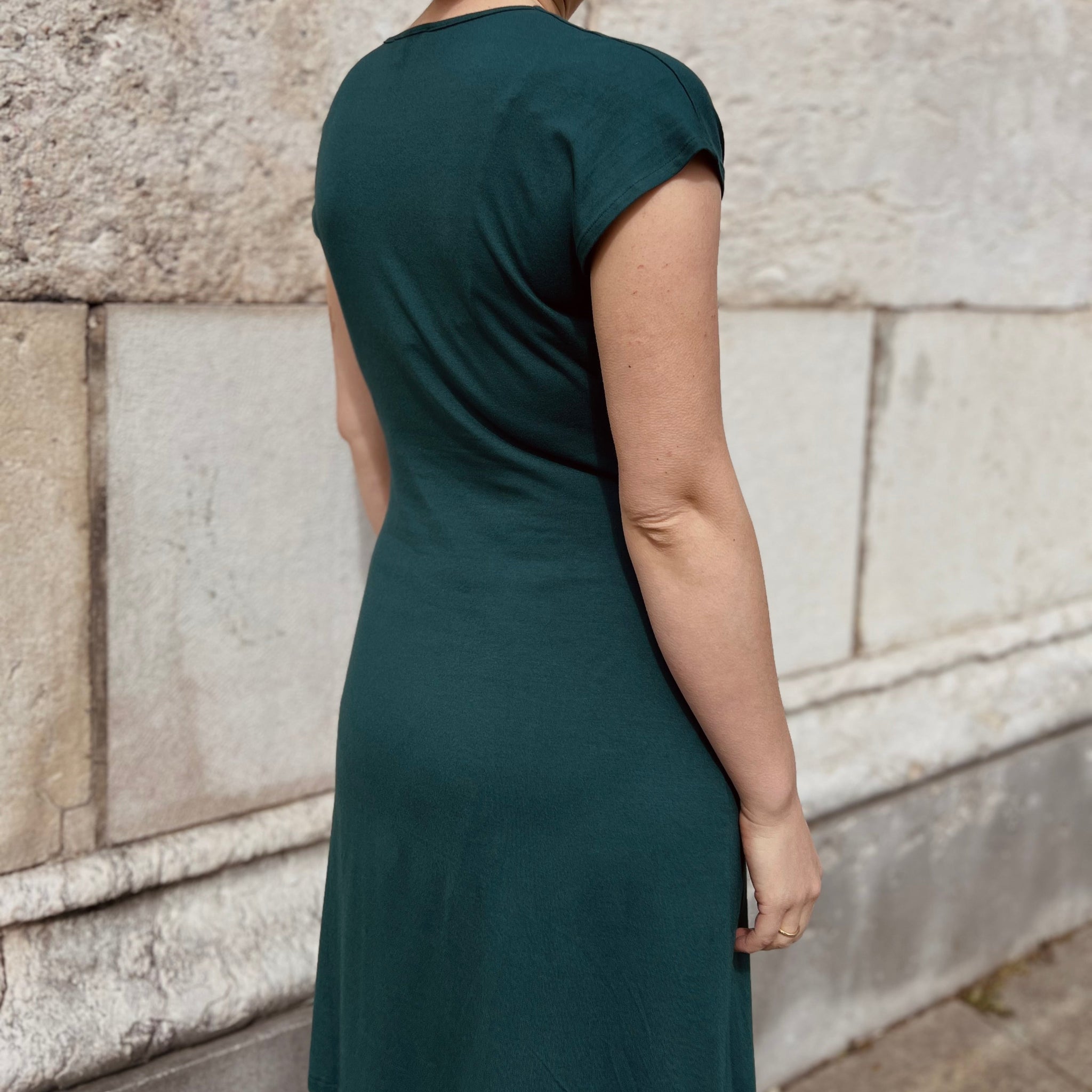 Eine Person in einem dunkelgrünen Kleid steht mit dem Rücken zur Kamera vor einer hellen Steinwand.