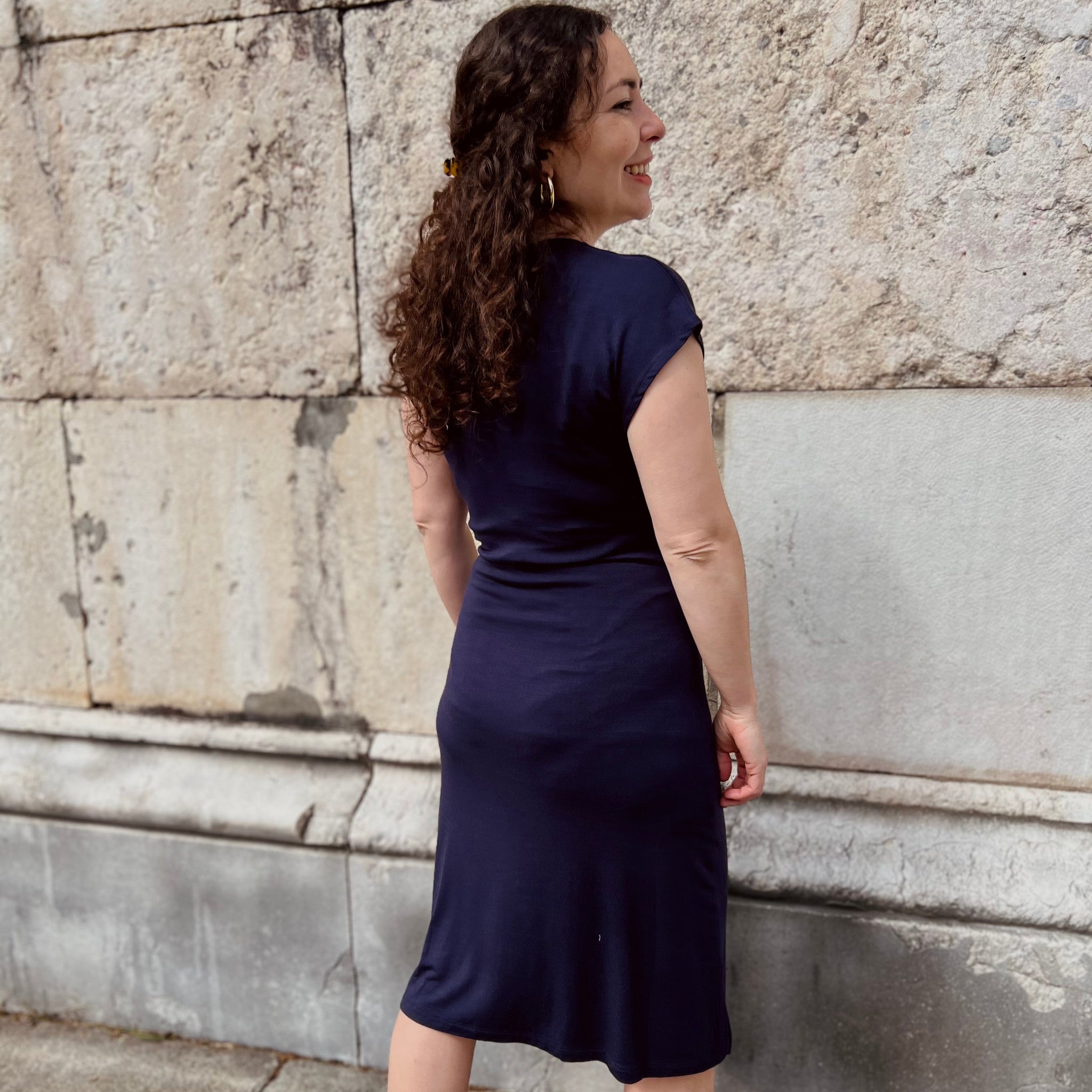 Eine Frau in einem dunkelblauen Kleid steht lächelnd vor einer hellen Steinwand.