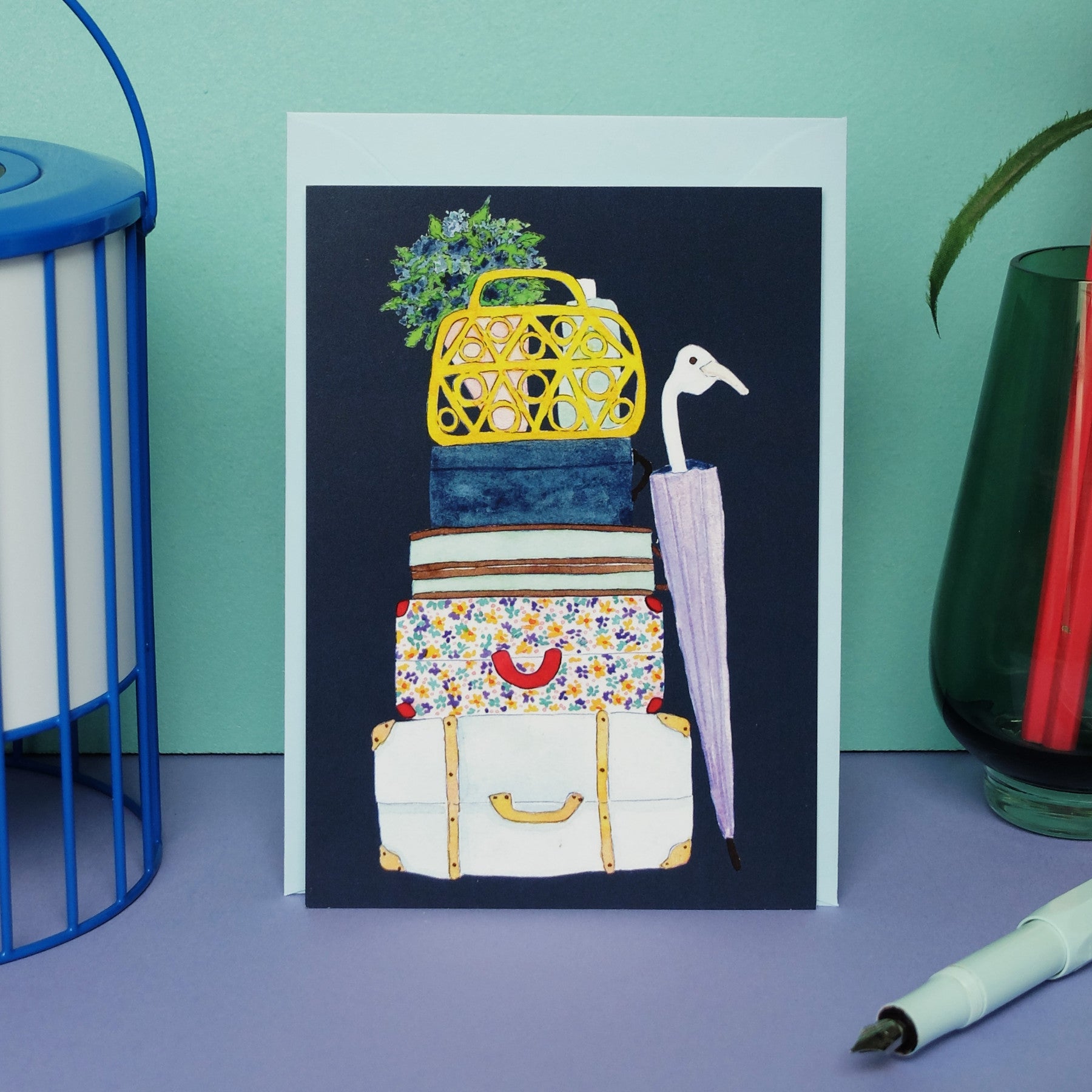 Eine Grußkarte mit einer Illustration von gestapelten Koffern, Büchern, einer blumengemusterten Tasche, einem gelben Korb mit Blumen und einem hellvioletten Regenschirm auf einer pastellfarbenen Fläche, daneben ein Stift und ein Glas mit Bleistiften.