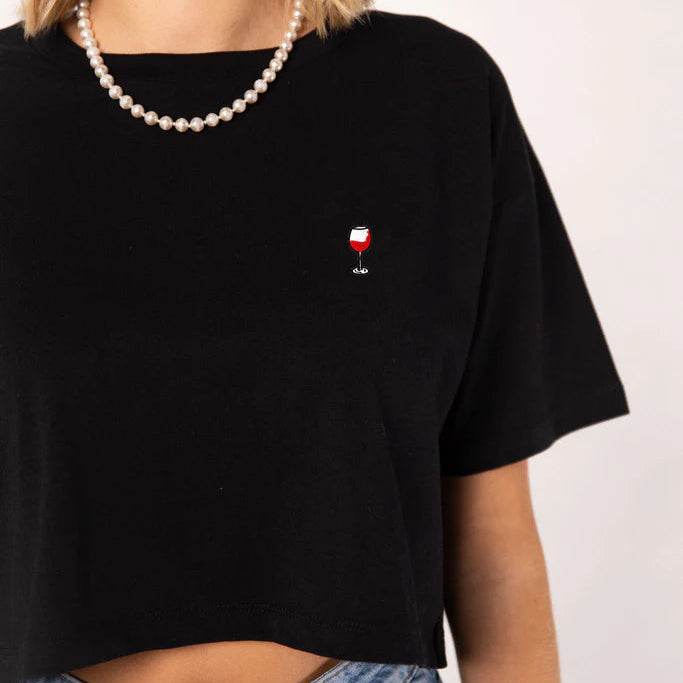 Eine Frau trägt ein schwarzes Crop-Top mit einem kleinen bestickten Glas Rotwein, eine Perlenkette und Blue Jeans. Das Bild ist so beschnitten, dass sie von den Schultern bis zur Taille zu sehen ist.