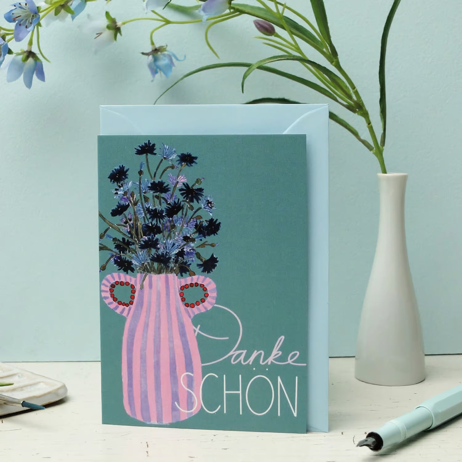 Eine Grußkarte mit einer Illustration von blauen und violetten Blumen in einer rosa gestreiften Vase, mit den Worten Danke Schön. Die Karte ist auf einem weißen Tisch neben einer Vase mit blauen Blumen und einem Stift aufgestellt.