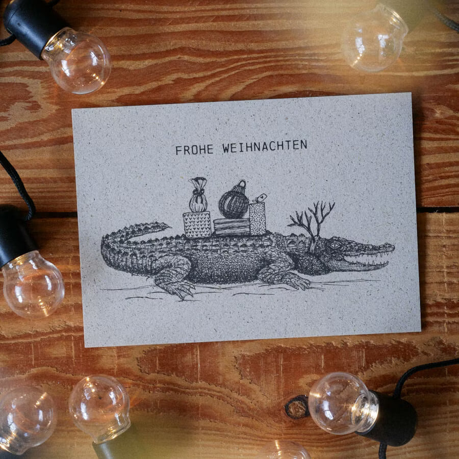 Weihnachtskarte mit einem gezeichneten Alligator, der Geschenke trägt, umgeben von Lichterketten auf Holztisch.