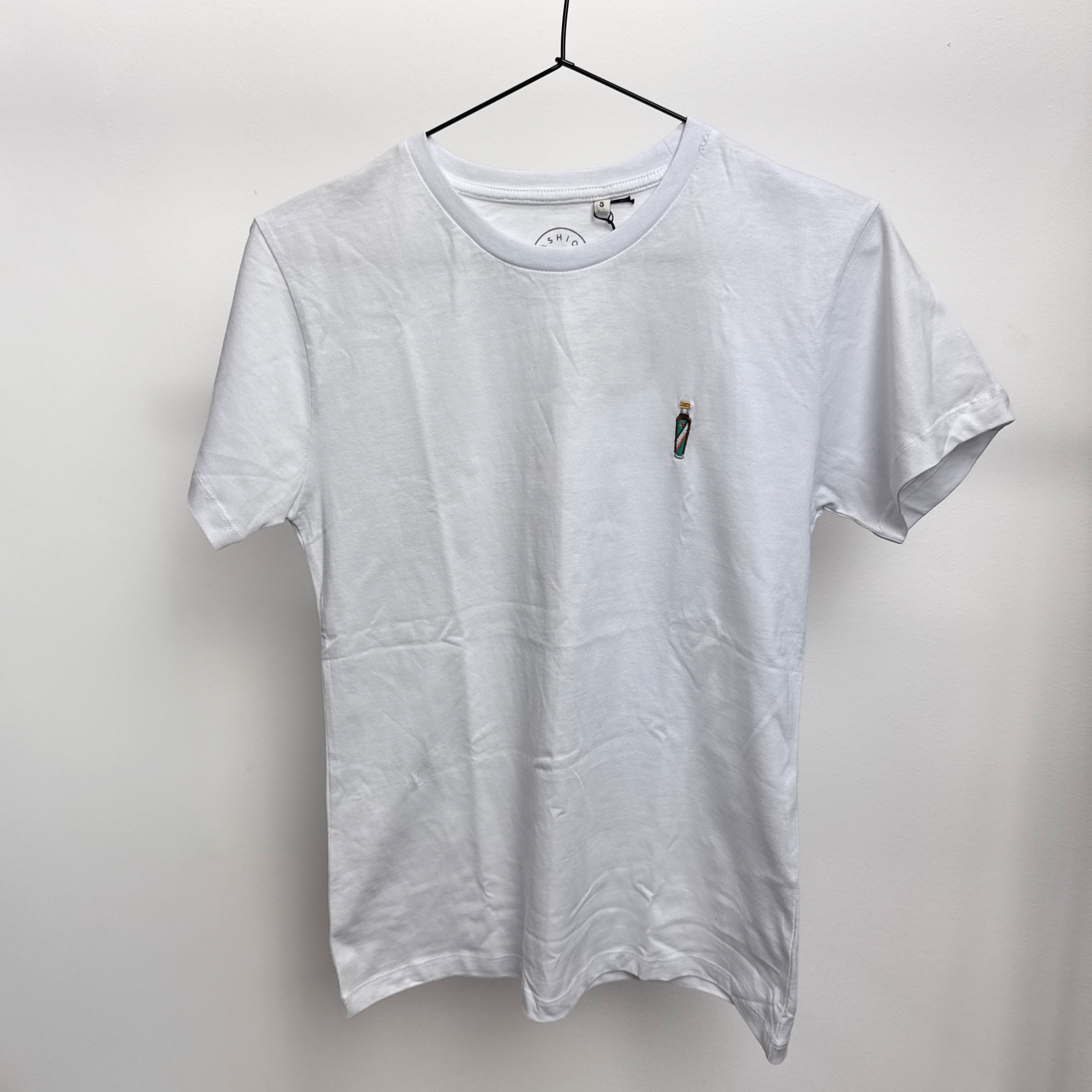 Ein schlichtes weißes T-Shirt hängt auf einem schwarzen Bügel vor einer weißen Wand. Auf dem T-Shirt ist auf der linken Brust ein kleines Flaschenmotiv aufgestickt.