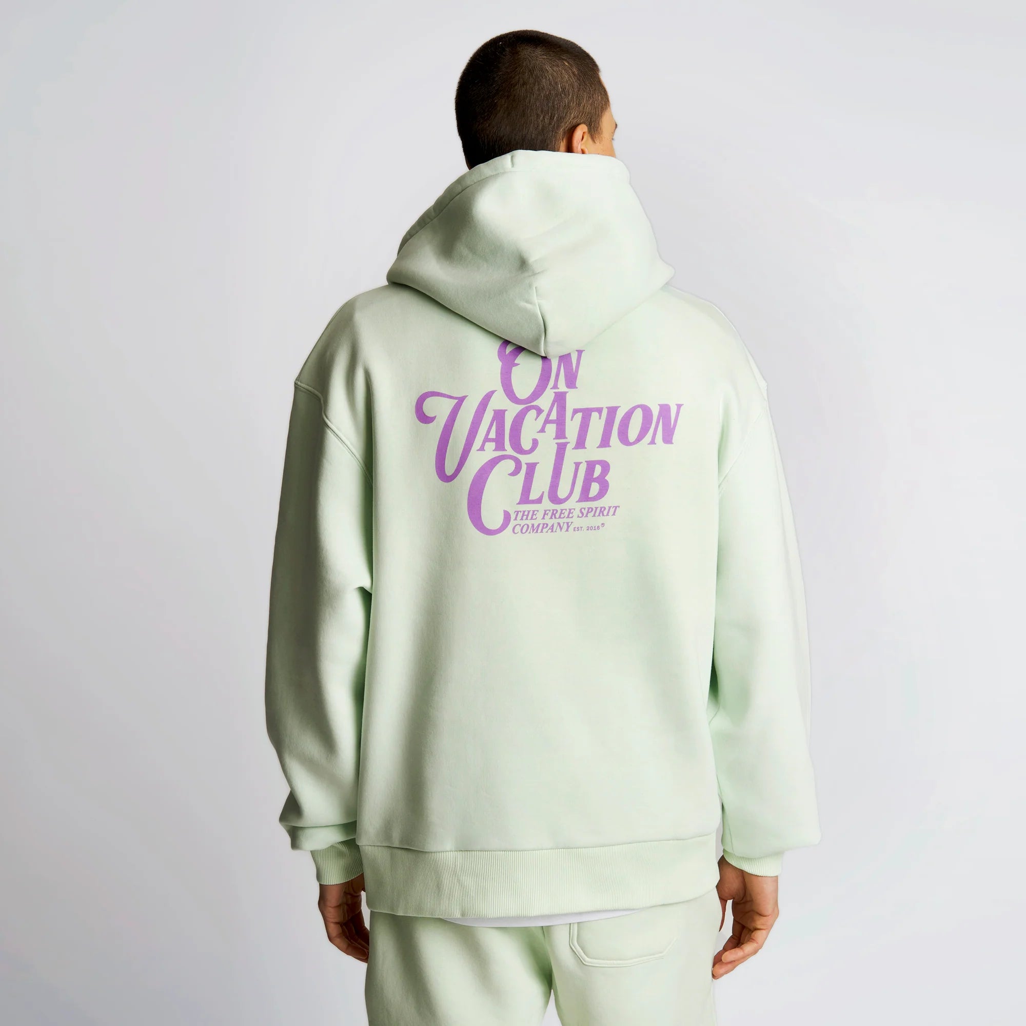 Person in mintgrünem Hoodie mit lila Schriftzug "ON VACATION CLUB" auf dem Rücken, steht mit dem Rücken zur Kamera.