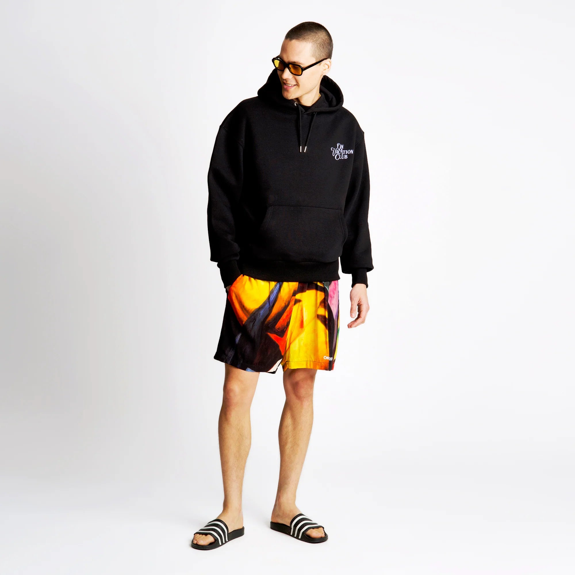 Mann in schwarzem Hoodie und bunt bedruckter Shorts steht auf weißem Hintergrund und trägt gestreifte Sandalen.