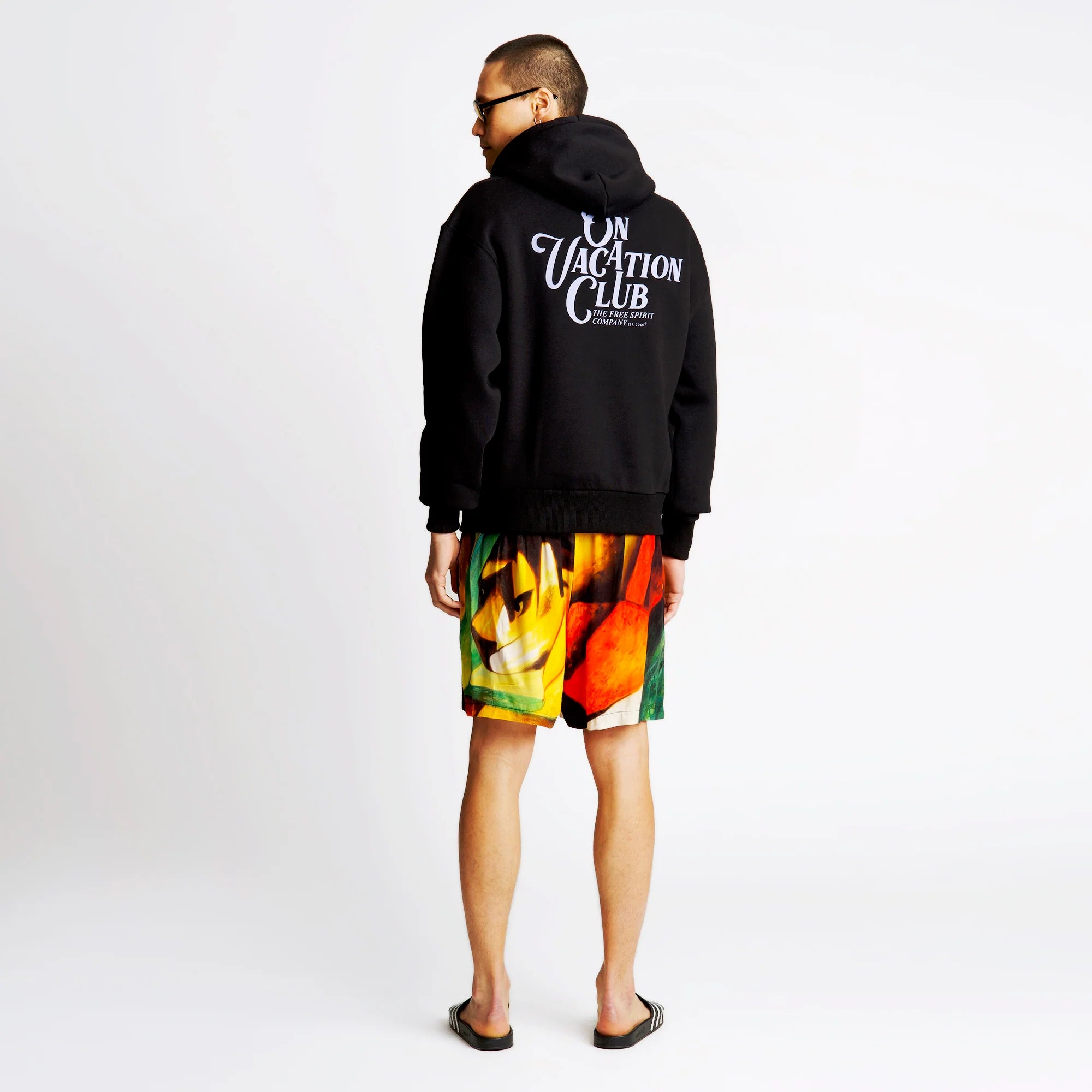 Person trägt einen schwarzen Hoodie mit "On Vacation Club"-Schriftzug und bunte Shorts, steht seitlich in Sandalen.