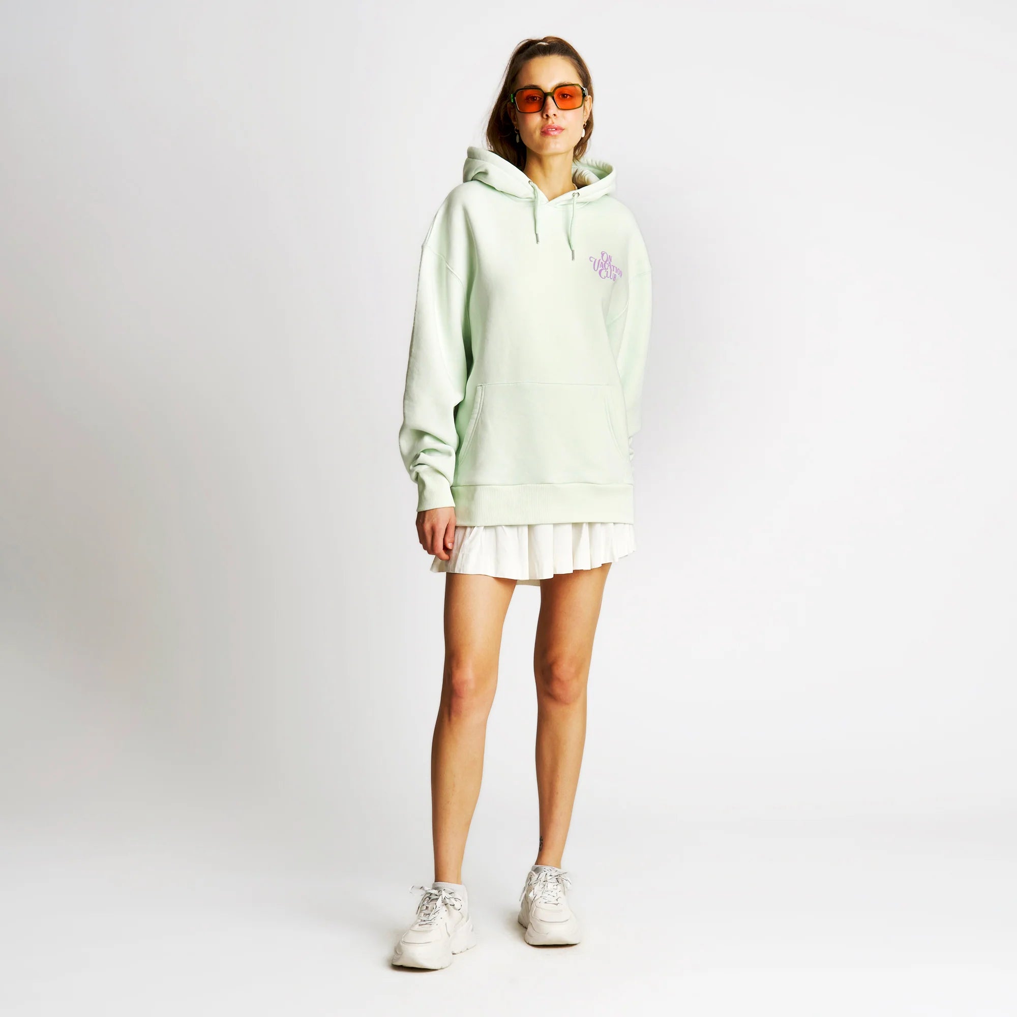 Frau in hellgrünem Hoodie, weißem Faltenrock und Sneakern, trägt eine orangefarbene Sonnenbrille, steht vor weißem Hintergrund.
