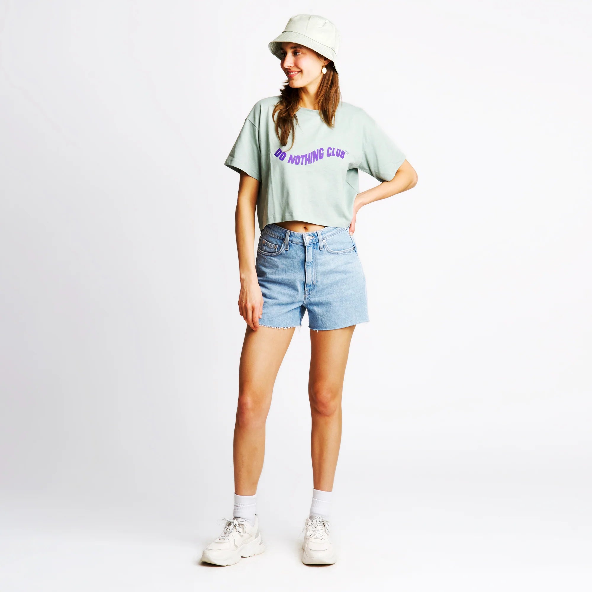 Frau trägt ein mintgrünes T-Shirt, Jeansshorts, weiße Schuhe und einen Hut vor weißem Hintergrund.