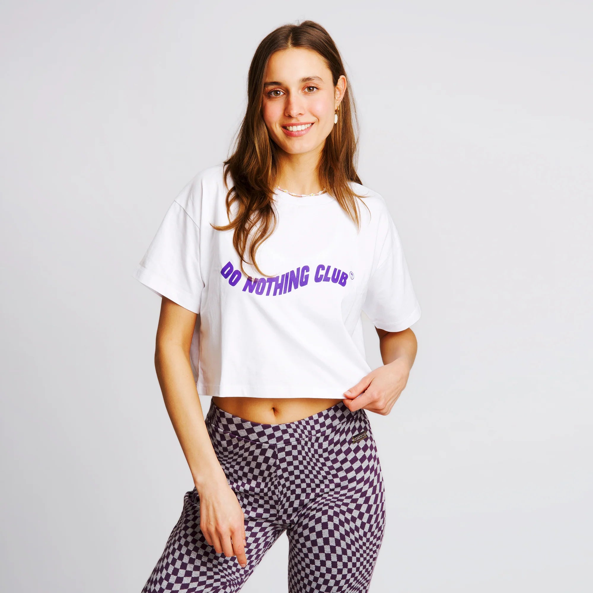 Frau in einem weißen T-Shirt mit "Do Nothing Club" Aufdruck und karierter Hose auf hellgrauem Hintergrund.