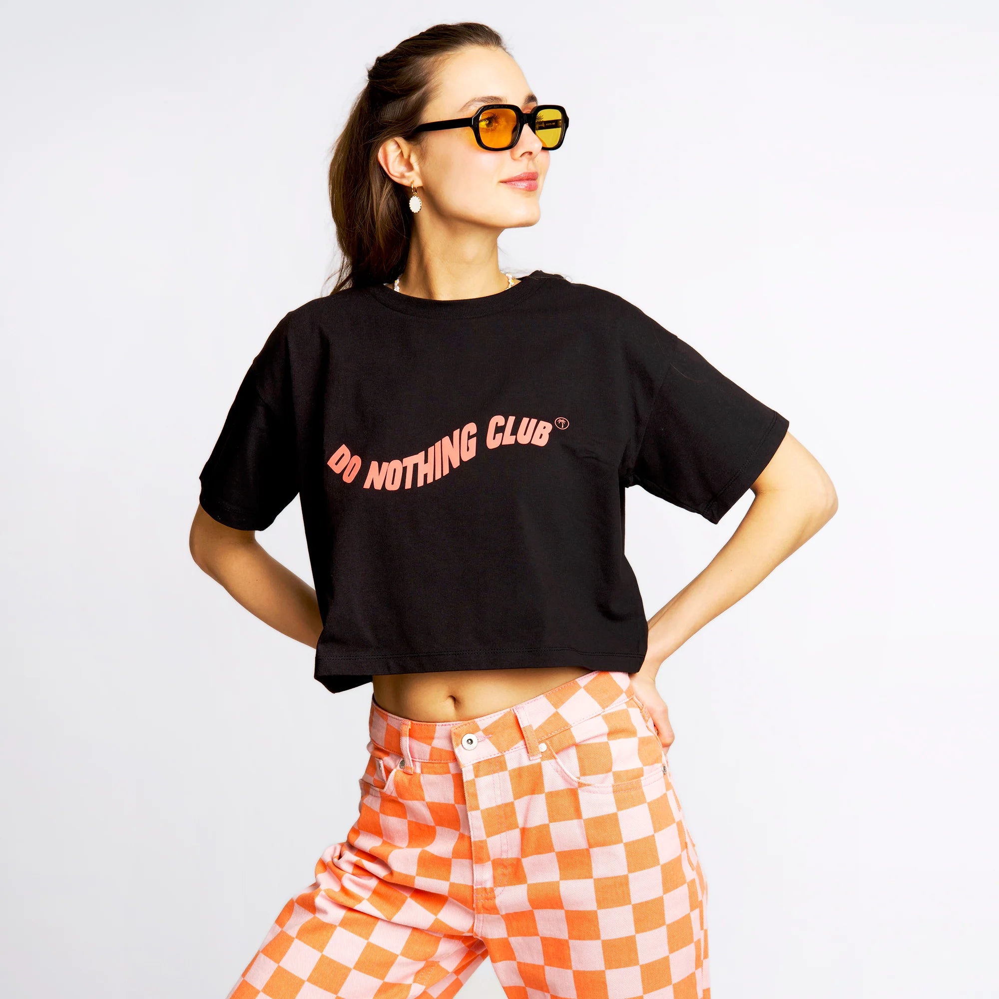 Frau mit Sonnenbrille trägt ein schwarzes T-Shirt mit der Aufschrift "Do Nothing Club" und karierte orange-weiße Hosen.
