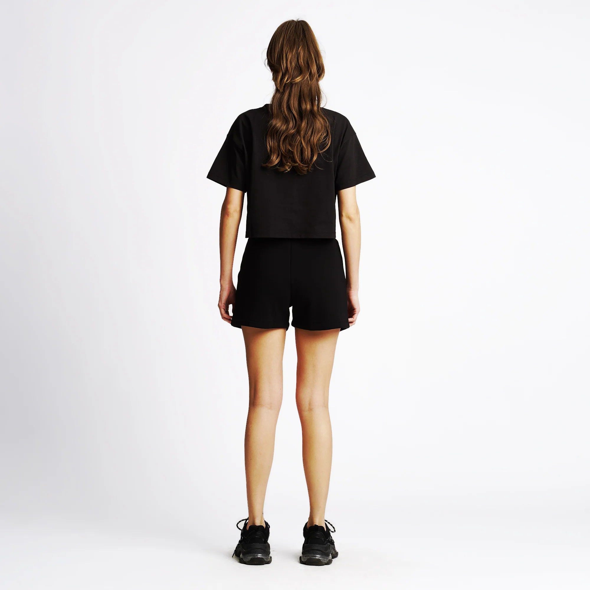 Person von hinten, trägt ein schwarzes T-Shirt, schwarze Shorts und schwarze Schuhe, steht vor einem weißen Hintergrund.