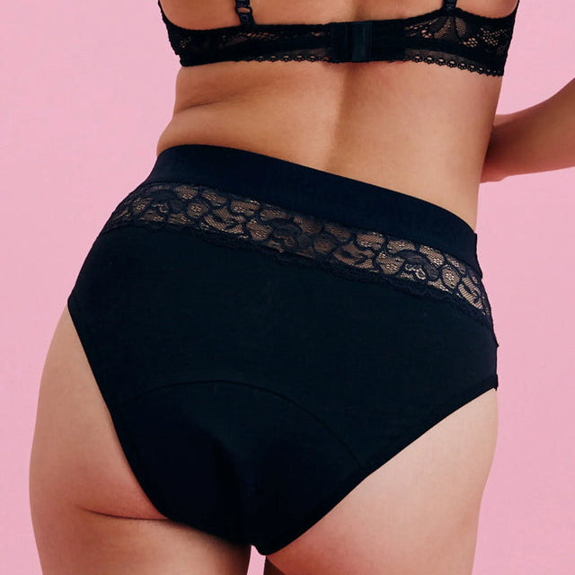 Eine Person wird von hinten gezeigt, die einen schwarzen Spitzen-BH und schwarze Unterwäsche mit hoher Taille und Spitzenverzierung trägt und vor einem rosa Hintergrund steht.