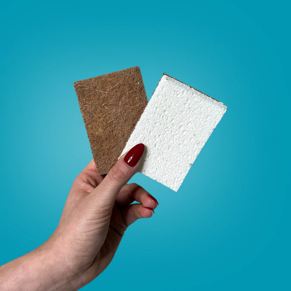 Eine Hand mit rot lackierten Nägeln hält zwei rechteckige Tafeln: eine braune und faserige, die andere weiße und strukturierte, vor blauem Hintergrund.