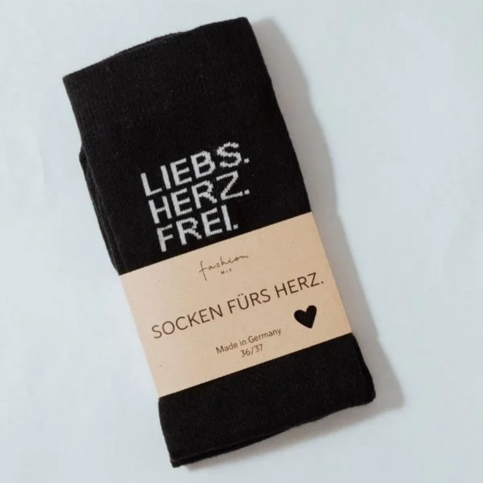 Ein Paar schwarze Socken mit weißer Aufschrift LIEBS. HERZ. FREI. Die Socken sind in ein beiges Etikett mit der Aufschrift SOCKEN FÜRS HERZ. und Made in Germany 36/37 mit einem kleinen Herzsymbol eingewickelt.