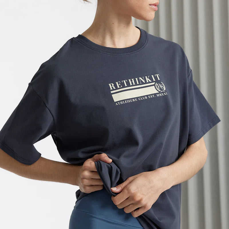Eine Frau in einem dunkelgrauen T-Shirt mit aufgedrucktem RETHINKIT-Text steht seitlich und hält das Hemd an der Hüfte. Sie hat ihr hellbraunes Haar zurückgesteckt und trägt blaue Leggings, mit neutralem Hintergrund und vertikalen Jalousien.