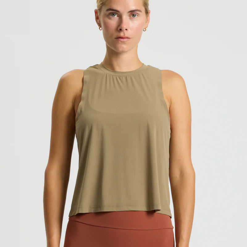 Eine Person, die ein ärmelloses, hellbraunes Tank-Top und rostfarbene Leggings mit hoher Taille trägt, steht mit dem Gesicht nach vorne vor einem schlichten hellen Hintergrund.