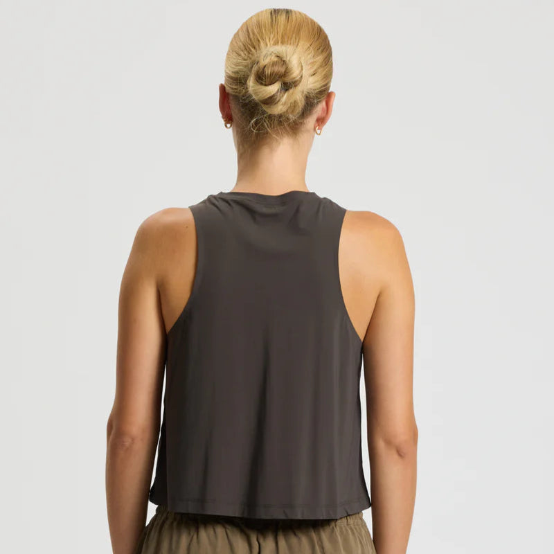 Eine Person mit blondem Haar in einem ordentlichen Dutt wird von hinten gezeigt. Sie trägt ein ärmelloses, locker sitzendes dunkelgraues Tanktop und eine olivgrüne Hose vor einem schlichten hellen Hintergrund.