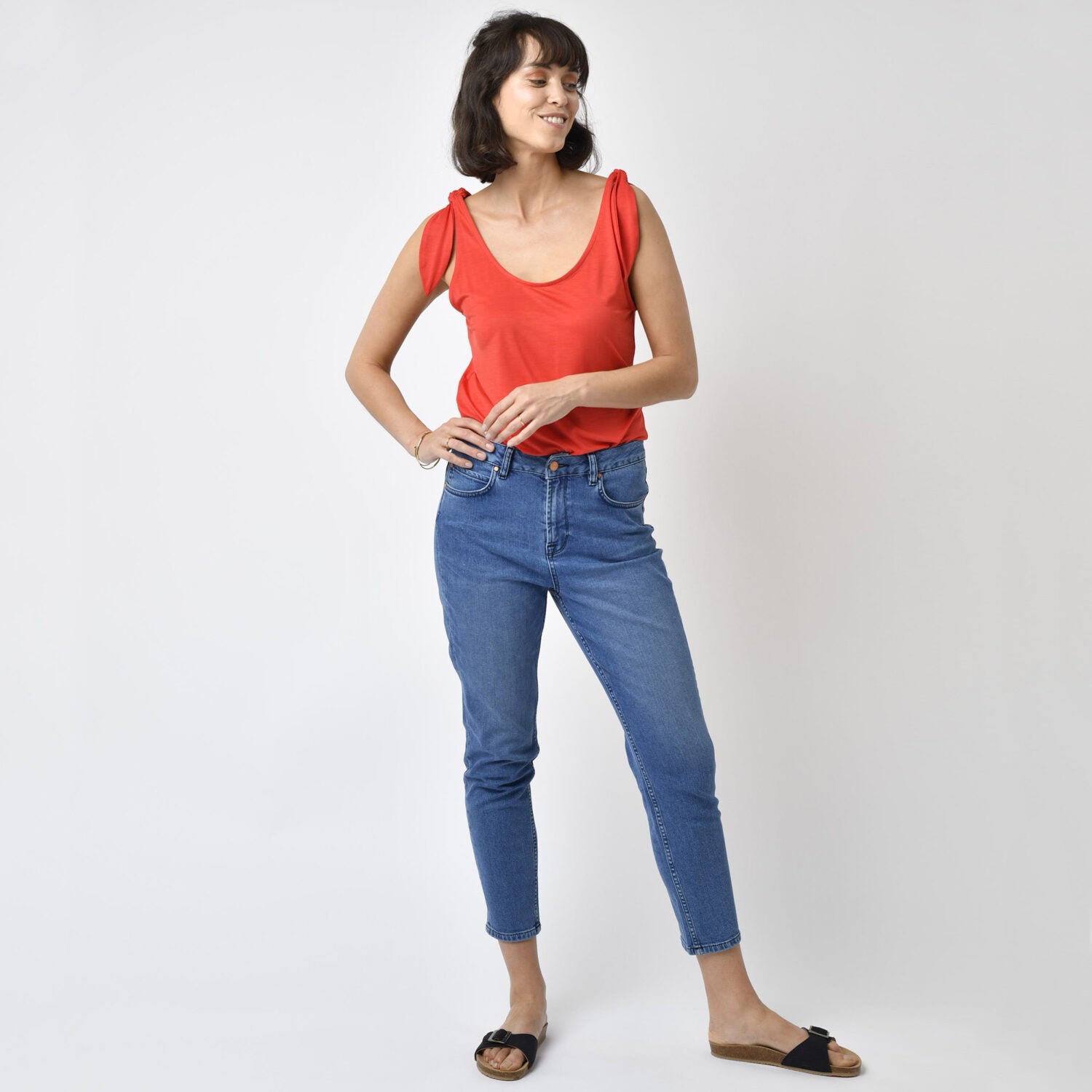 Frau in rotem Top und blauer Jeans posiert lächelnd vor neutralem Hintergrund.