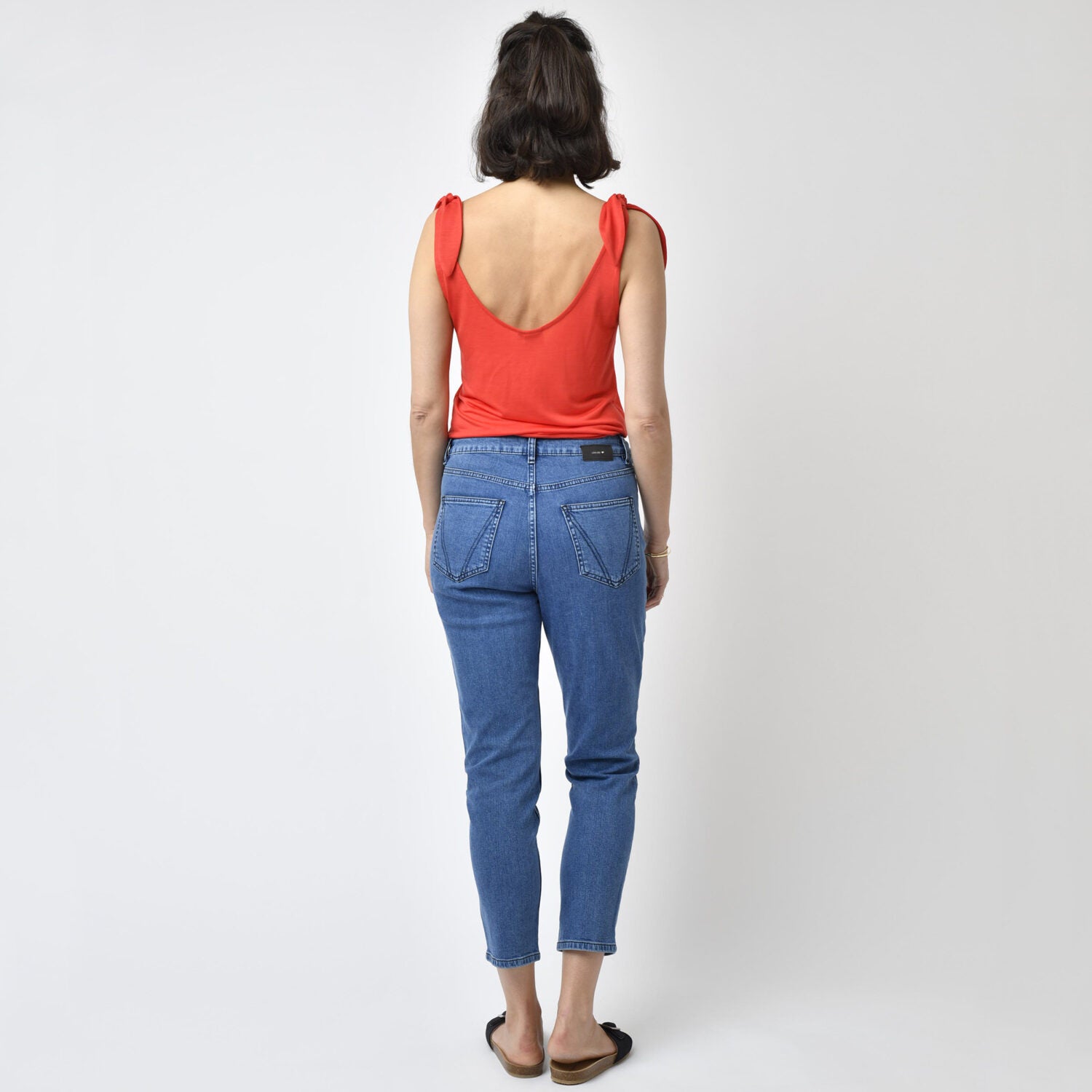 Eine Frau in einem roten Oberteil und blauer Jeans steht mit dem Rücken zur Kamera vor einem neutralen Hintergrund.