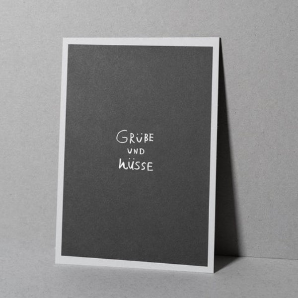 Eine schwarze Grußkarte mit weißem Rand steht aufrecht. In der Mitte steht handgeschriebener weißer Text: "Grüße und Küsse". Der Hintergrund ist eine schlichte graue Fläche.