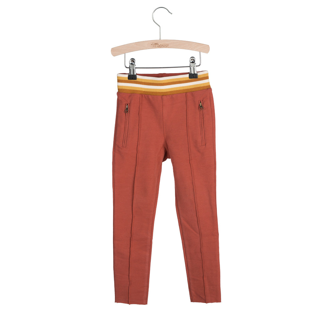 Rostfarbene Kinderhose mit Reißverschlüssen an der Vorderseite und einem gelb, weiß und rostfarben gestreiften Gummibund, aufgehängt an einem Holzbügel vor weißem Hintergrund.