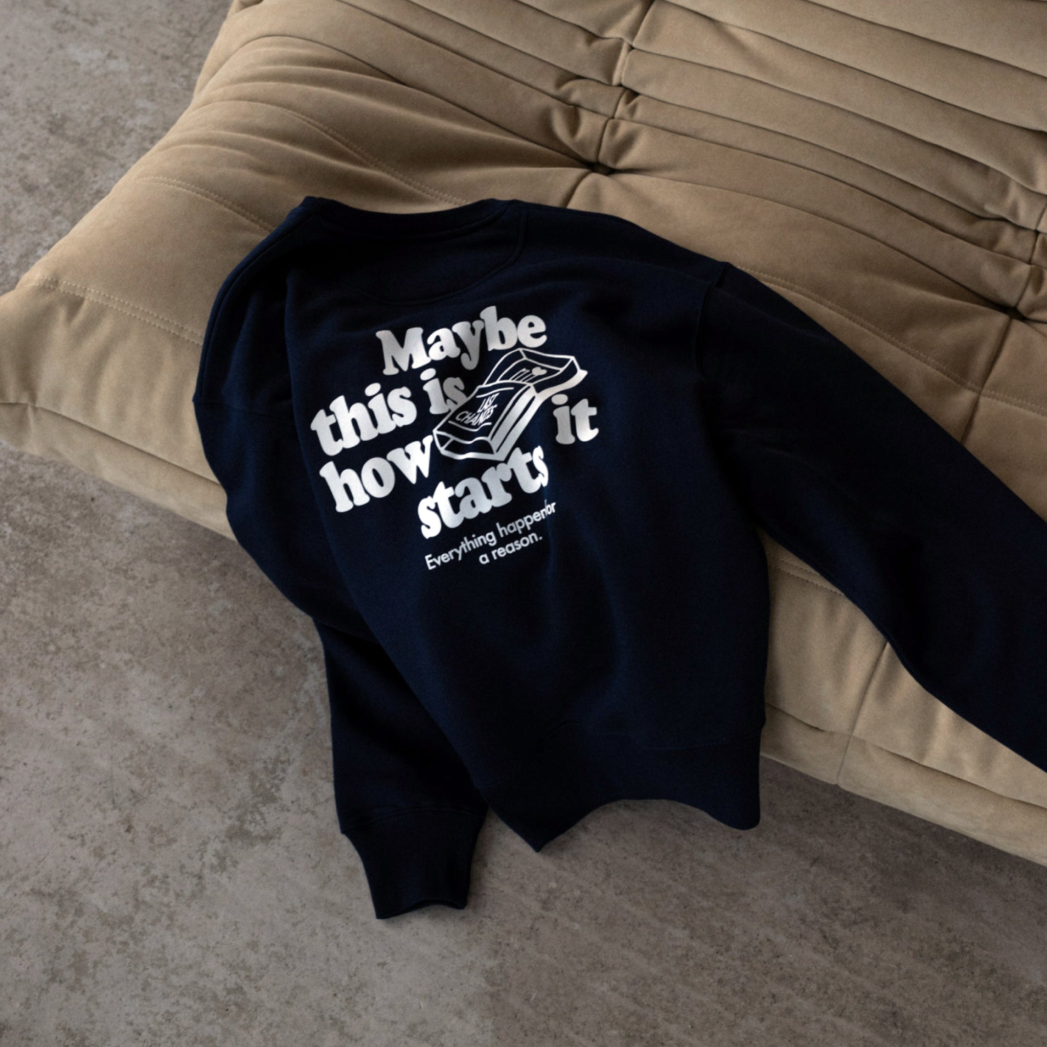 Dunkelblauer Pullover mit weißem Text und Grafik liegt auf einem beigen, gesteppten Kissen auf einem Betonboden.