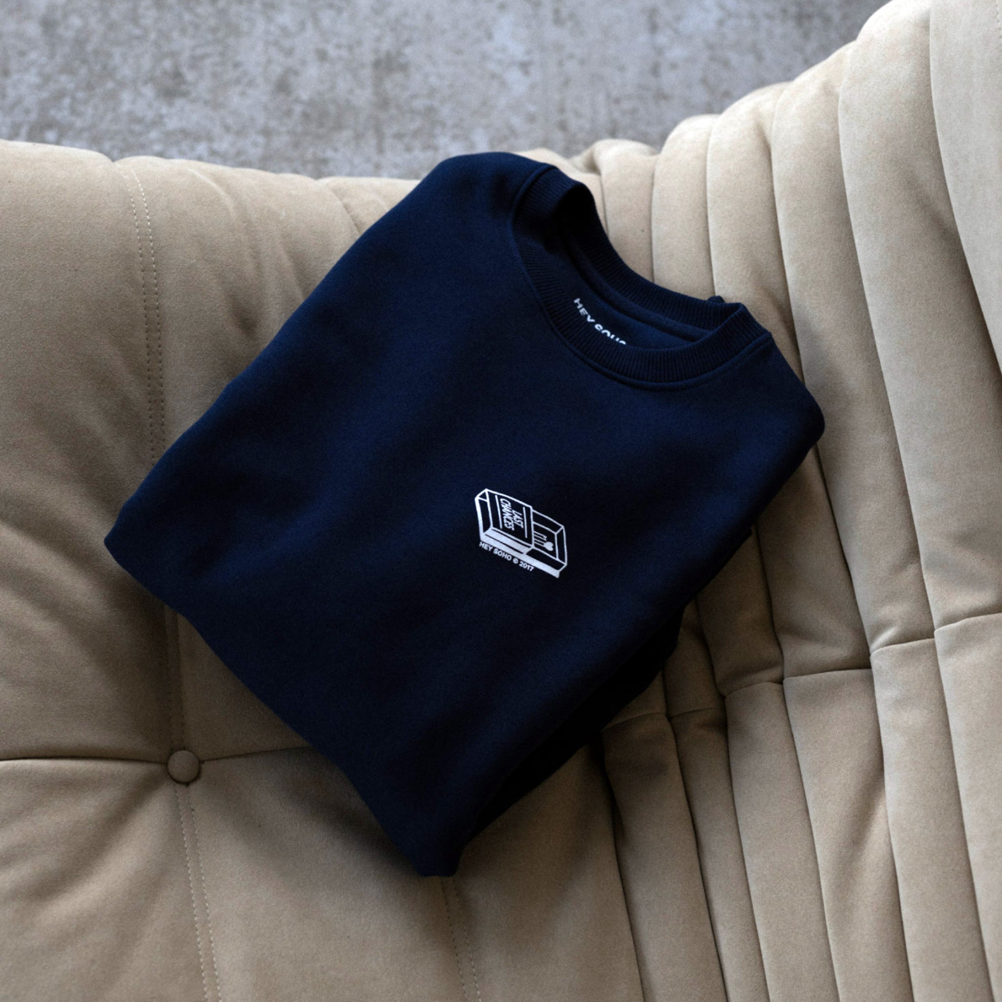 Gefalteter dunkelblauer Pullover mit kleinem weißen Logo, liegt auf einem beigefarbenen Polstermöbel.
