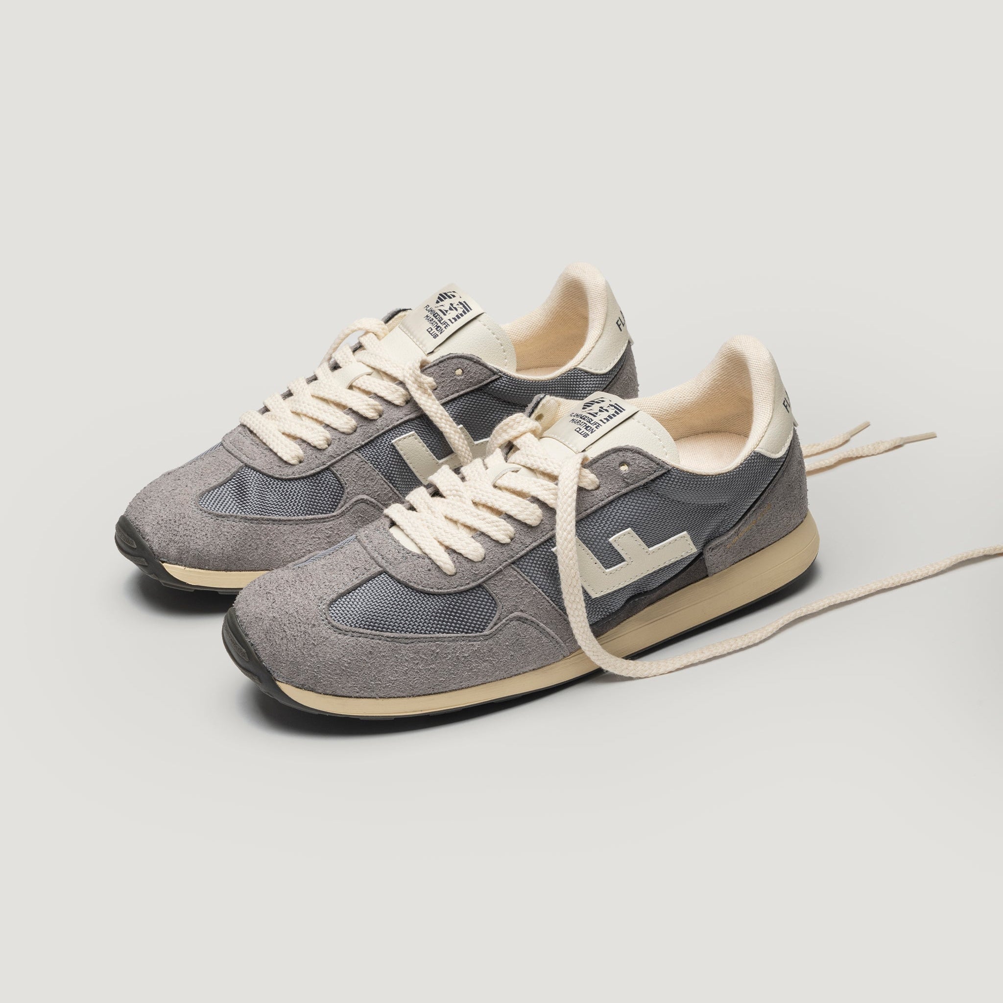 Ein Paar graue und weiße Turnschuhe im Retrostil mit cremefarbenen Schnürsenkeln, die nebeneinander auf einem schlichten hellen Hintergrund abgebildet sind. Ein Schnürsenkel ist locker vor den Schuhen verlängert.