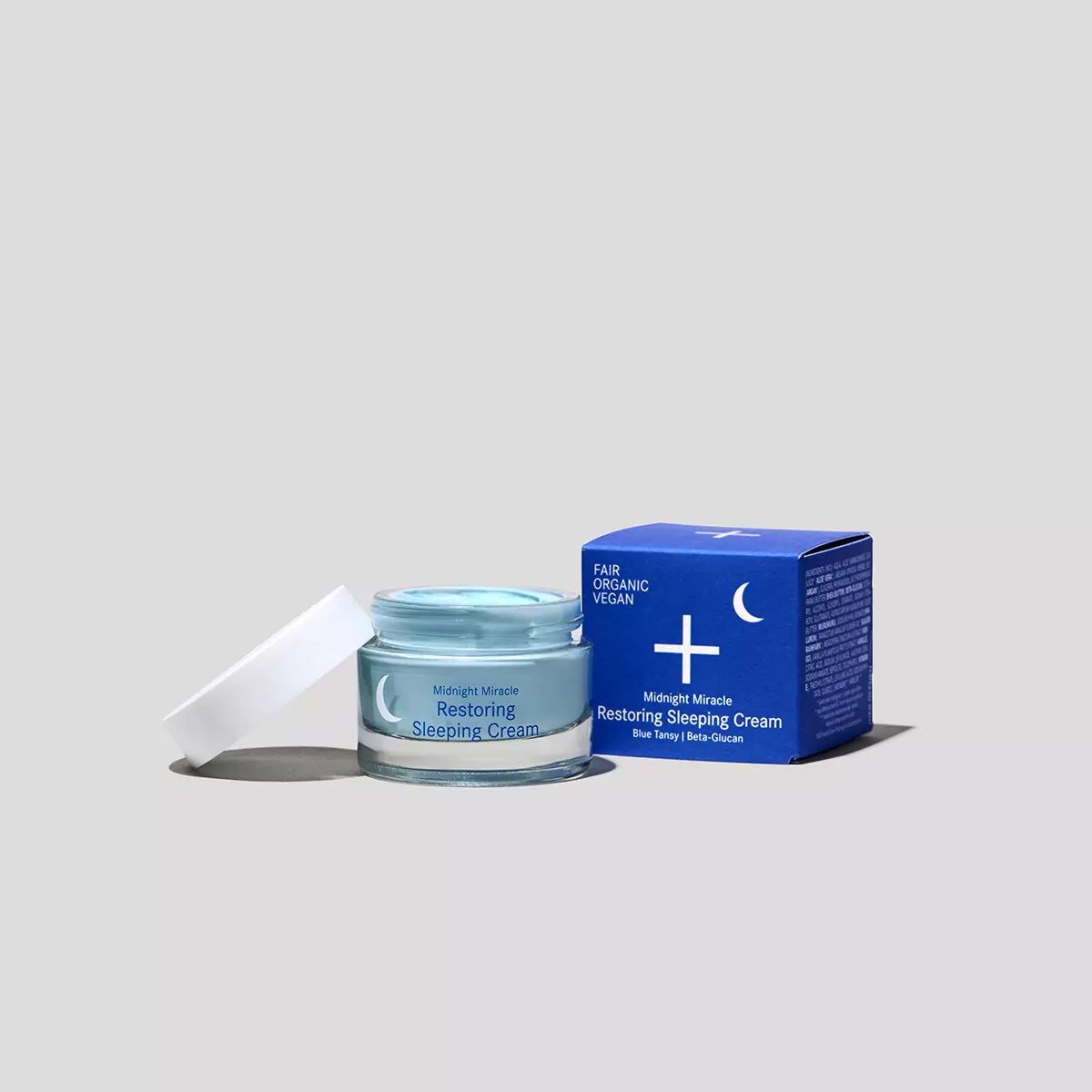 Ein Tiegel der Midnight Miracle Restoring Sleeping Cream mit geöffnetem weißen Deckel, daneben eine blaue Produktverpackung mit einem Halbmond und dem Hinweis, dass sie fair, biologisch und vegan ist, alles vor einem grauen Hintergrund.