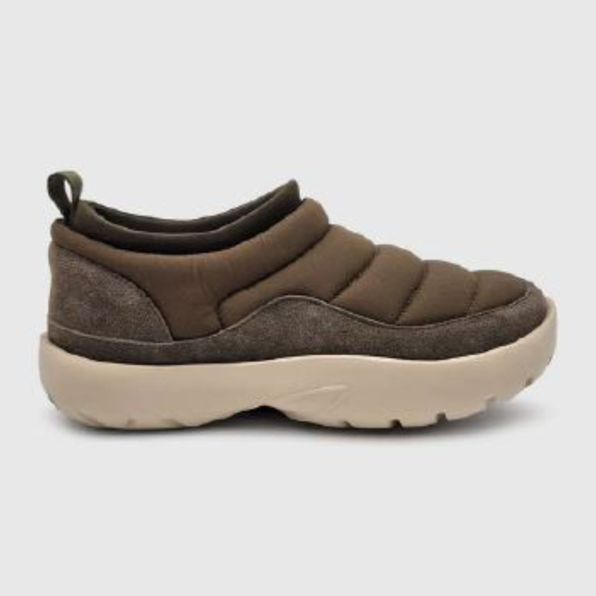 Ein brauner Slipper mit gestepptem, gepolstertem Obermaterial und einer dicken, hellbeigen Gummisohle, von der Seite gesehen vor einem schlichten grauen Hintergrund.