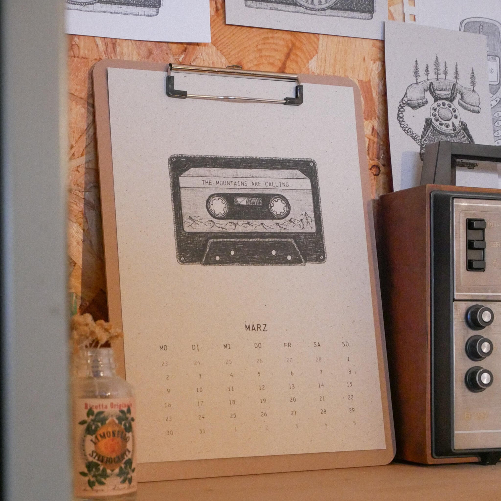 Auf einem Schreibtisch liegt ein Kalender mit dem Monat März. Auf seiner Seite ist eine Audiokassette mit der Aufschrift THE MOUNTAINS ARE CALLING abgebildet. In der Nähe stehen ein Radio, eine Glasflasche und Schwarz-Weiß-Zeichnungen an einer Holzwand.