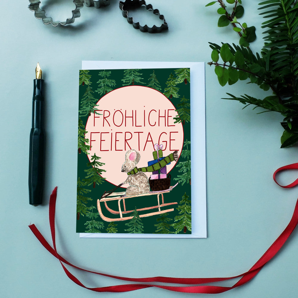 Eine Weihnachtskarte mit der Zeichnung einer Maus auf einem Schlitten mit Geschenken, umgeben von Bäumen und der Aufschrift FRÖHLICHE FEIERTAGE. Die Karte liegt neben einem Füllfederhalter, Band, Grünzeug und Ausstechformen.