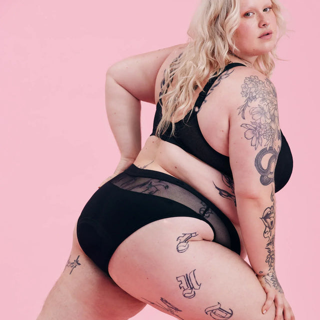 Eine Person mit langen blonden Haaren posiert selbstbewusst in schwarzen Dessous vor einem rosafarbenen Hintergrund und zeigt mehrere sichtbare Tätowierungen an Armen, Oberkörper und Beinen.