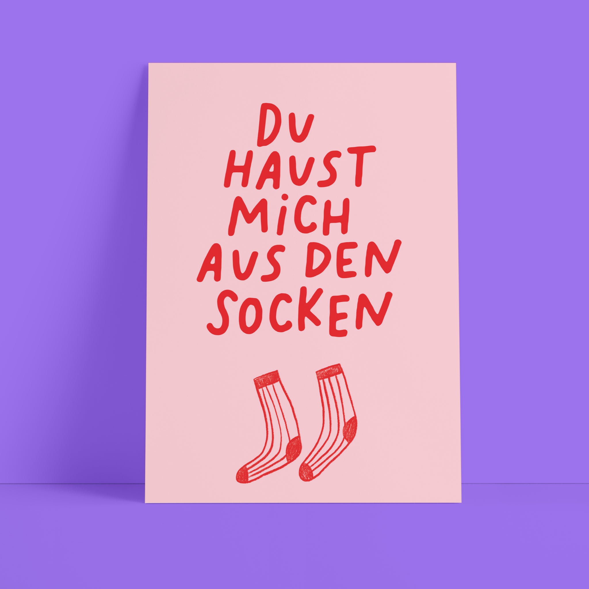 Eine rosafarbene Karte mit rotem handgeschriebenem deutschem Text Du haust mich aus den Socken über einer Zeichnung von zwei roten Socken, die aufrecht vor einem lila Hintergrund stehen.