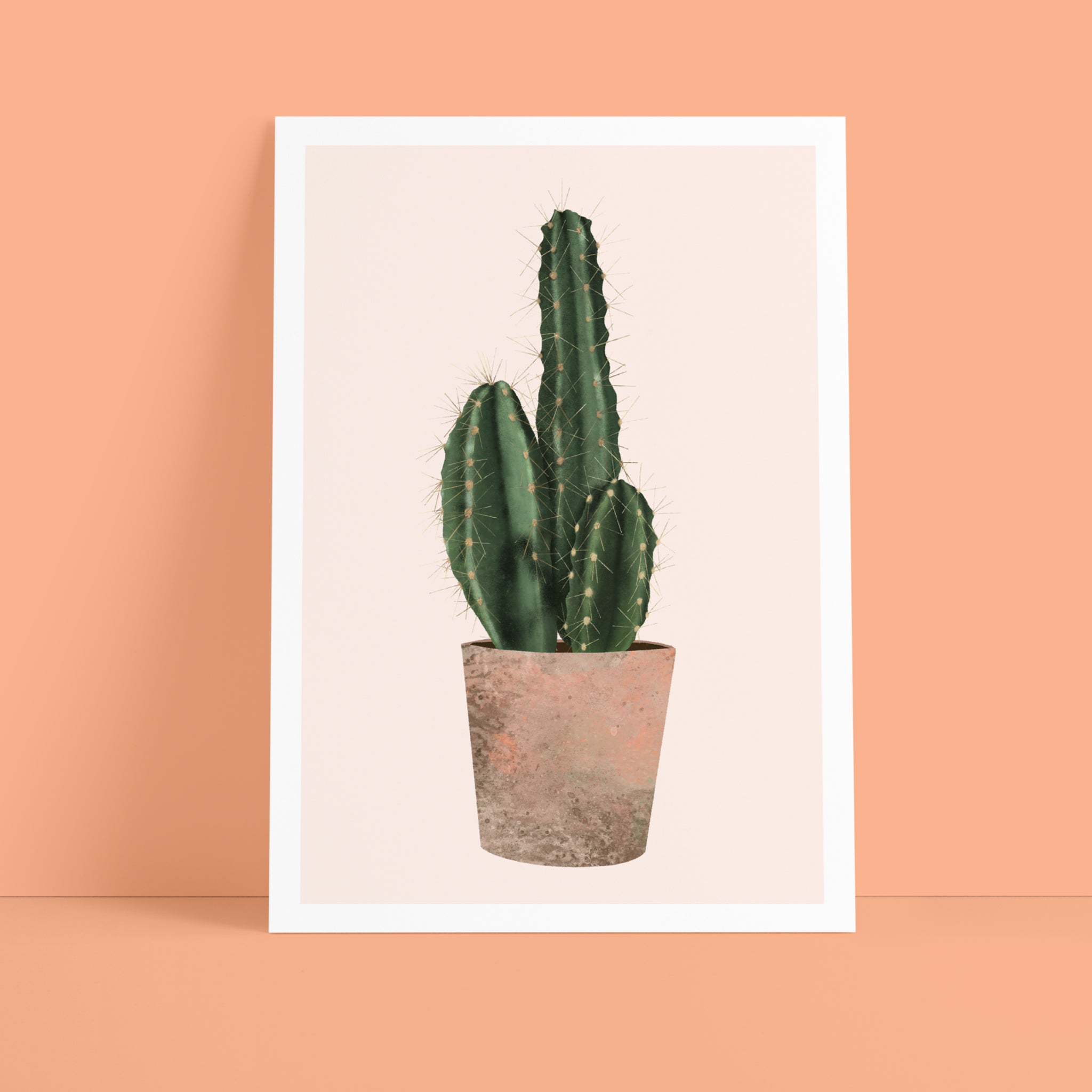 Ein minimalistischer Druck eines grünen, getopften Kaktus wird vor einem sanften pfirsichfarbenen Hintergrund gezeigt. Der Kaktus steht in der Mitte eines weißen Rahmens, wobei das Kunstwerk leicht an die Wand gelehnt ist.