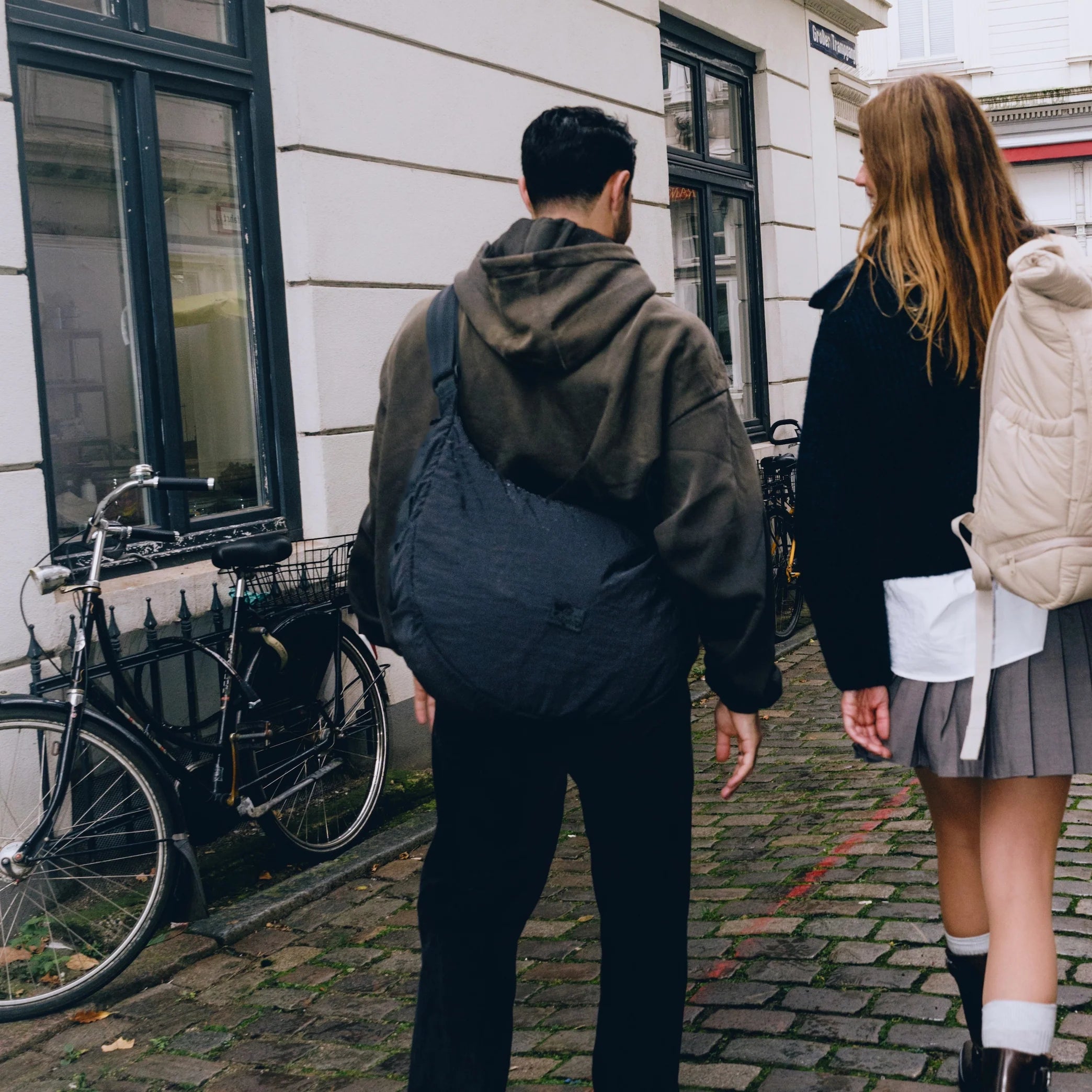 Zwei Personen gehen auf einer Straße mit Kopfsteinpflaster neben geparkten Fahrrädern und einem weißen Gebäude. Die eine trägt einen Kapuzenpulli und eine Umhängetasche, die andere trägt einen Rock, einen schwarzen Pullover und einen Rucksack.