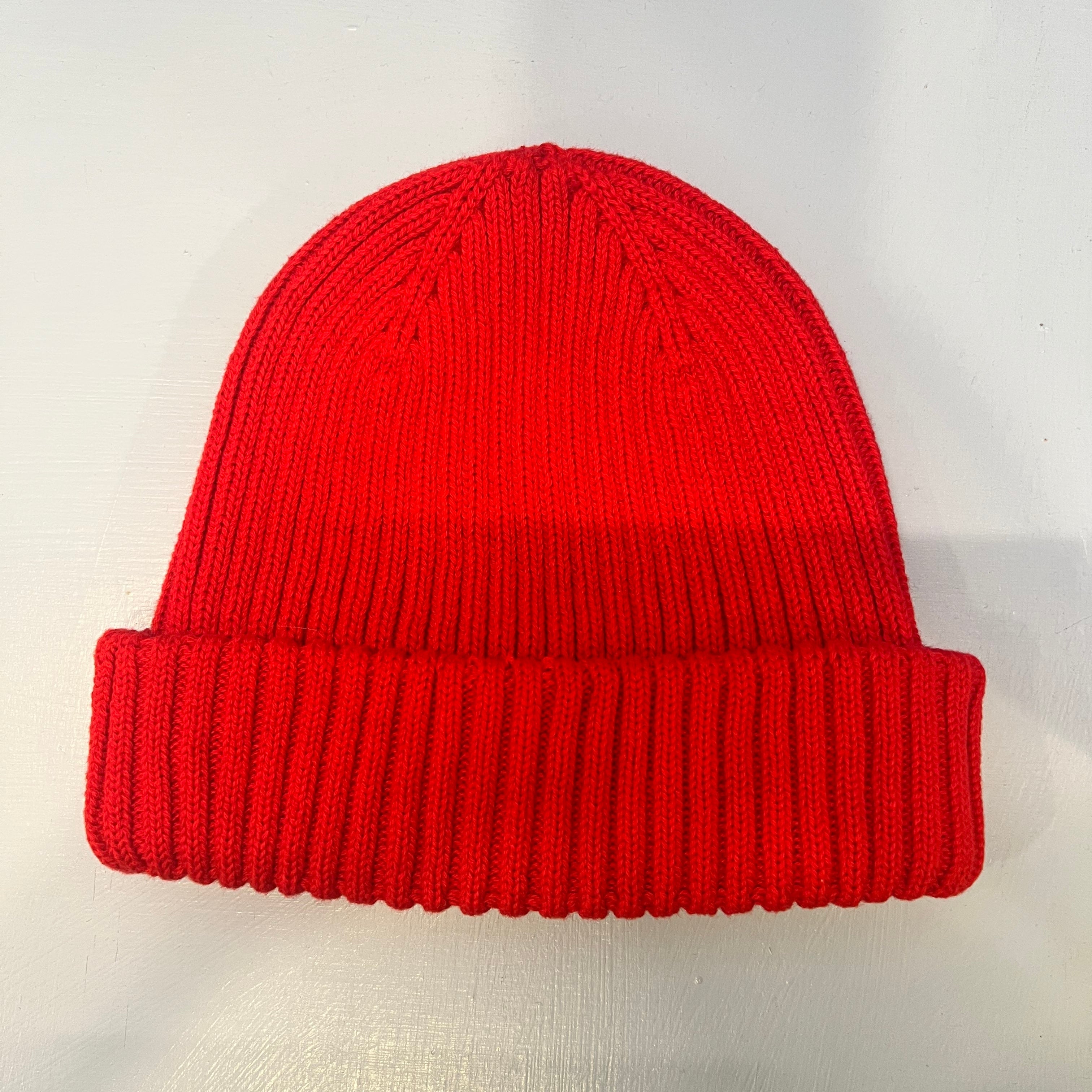 Eine leuchtend rote Strickmütze mit umgeschlagener Krempe wird auf eine hellgraue Fläche gelegt.