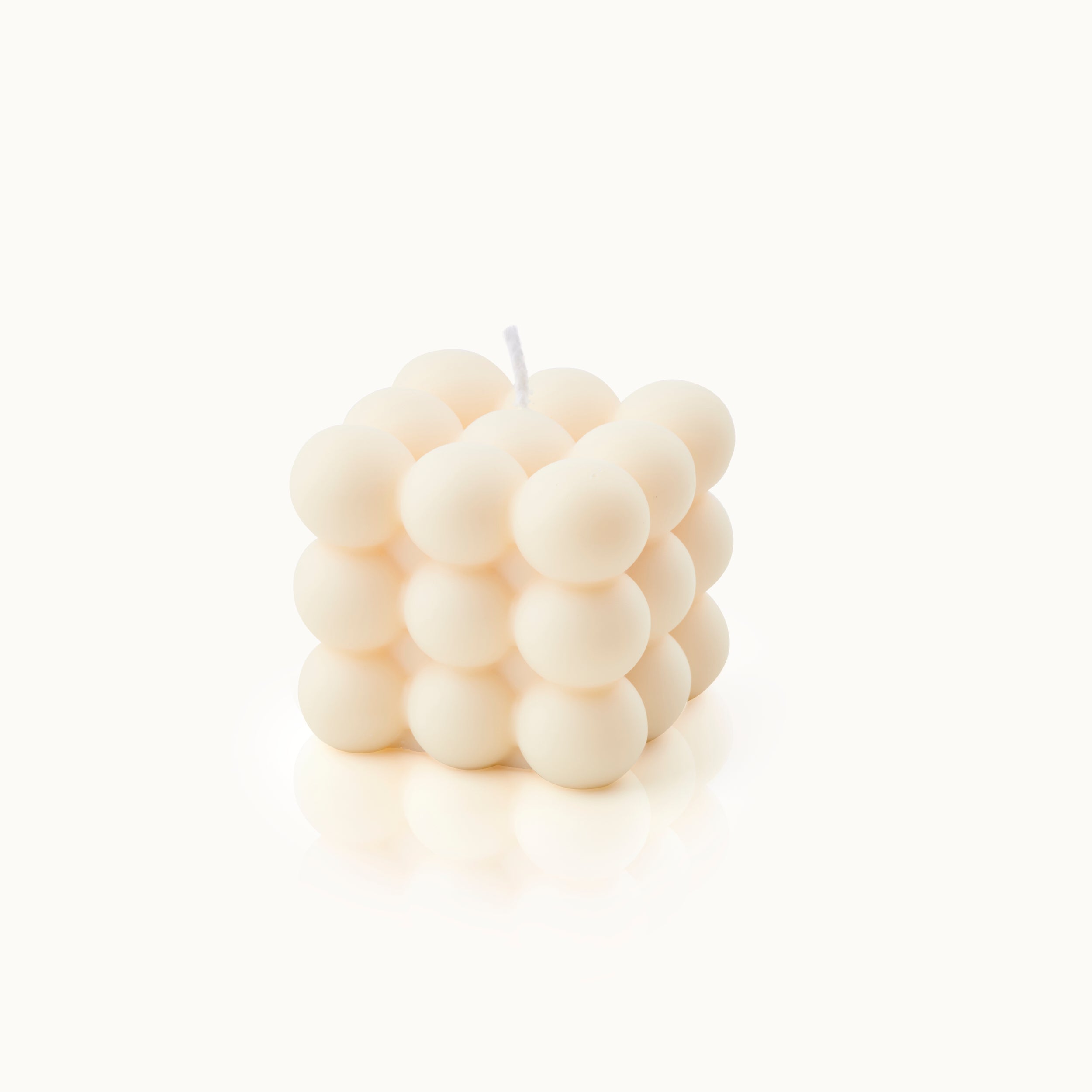 Eine cremefarbene Kerze in Form eines Würfels, dessen Oberfläche aus runden, blasenartigen Kugeln besteht. Die Kerze hat einen weißen Docht an der Spitze und steht vor einem schlichten weißen Hintergrund.