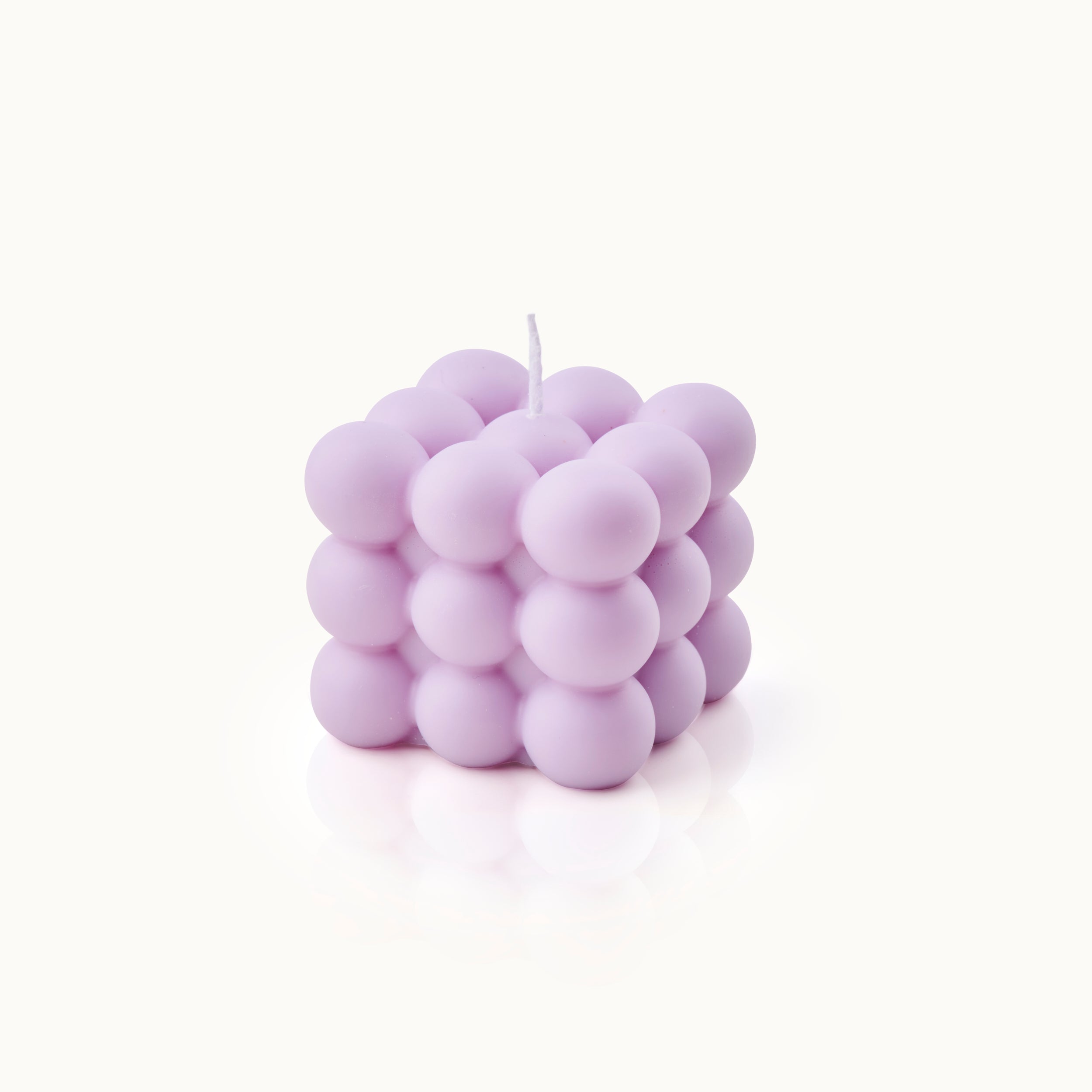 Eine lavendelfarbene Kerze in Form eines Würfels, bestehend aus kleinen runden Kugeln, die aneinander gereiht sind, mit einem weißen Docht obenauf, vor einem schlichten weißen Hintergrund.