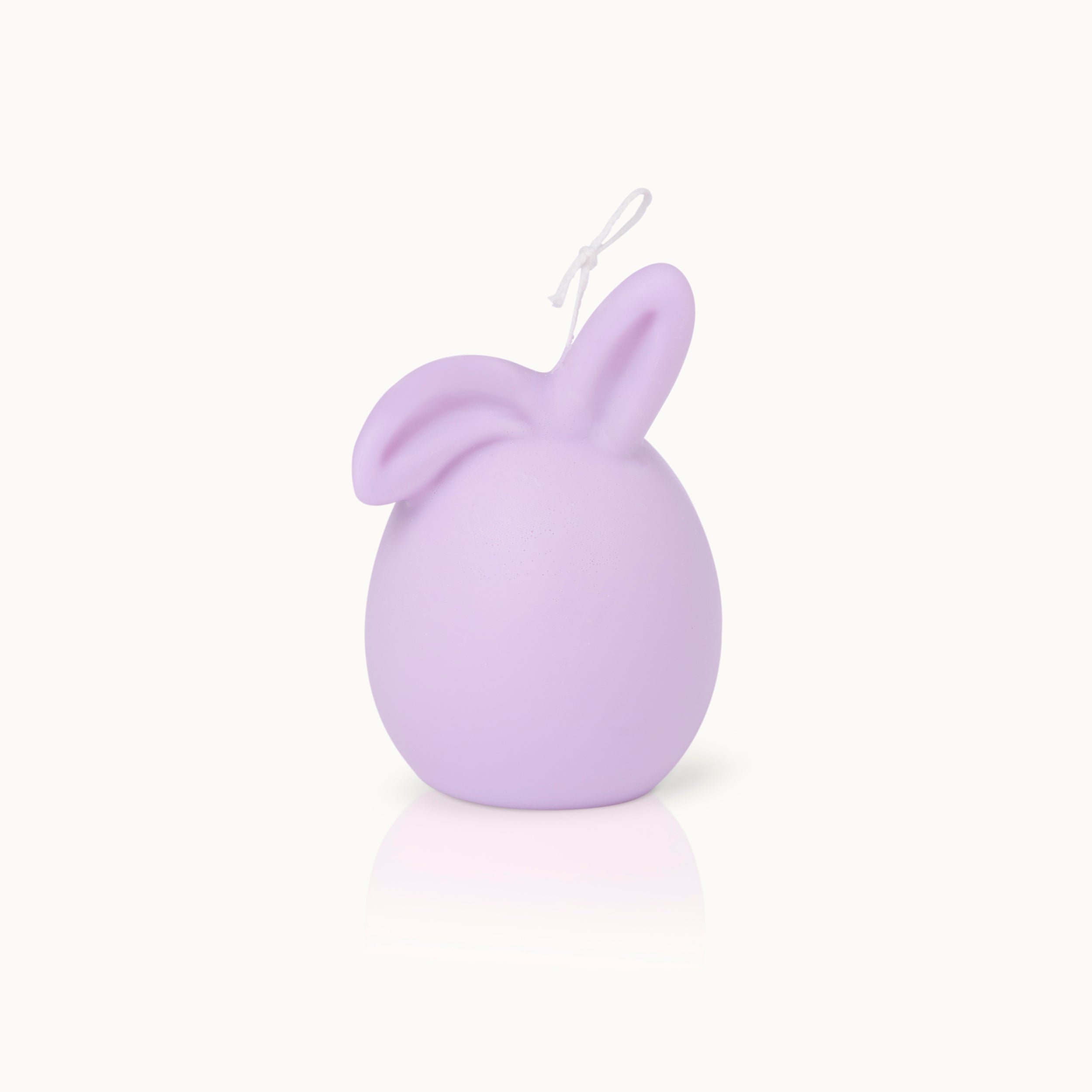 Eine pastellviolette, eiförmige Kerze mit zwei Hasenohren an der Spitze und einem weißen Docht vor einem schlichten weißen Hintergrund.