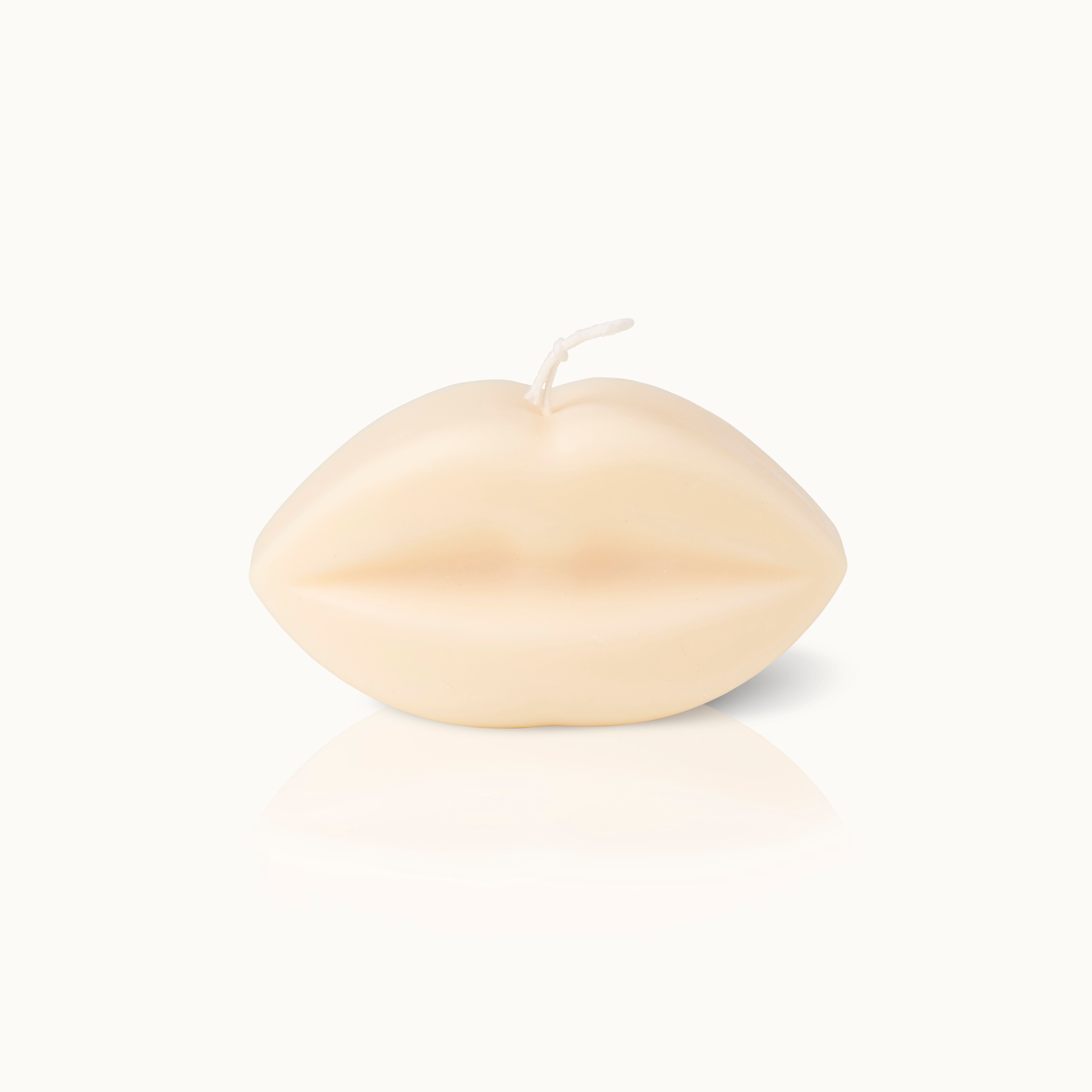 Eine cremefarbene Kerze in Form eines Lippenpaares steht auf einer weißen Fläche, mit einem kurzen Docht an der Spitze.