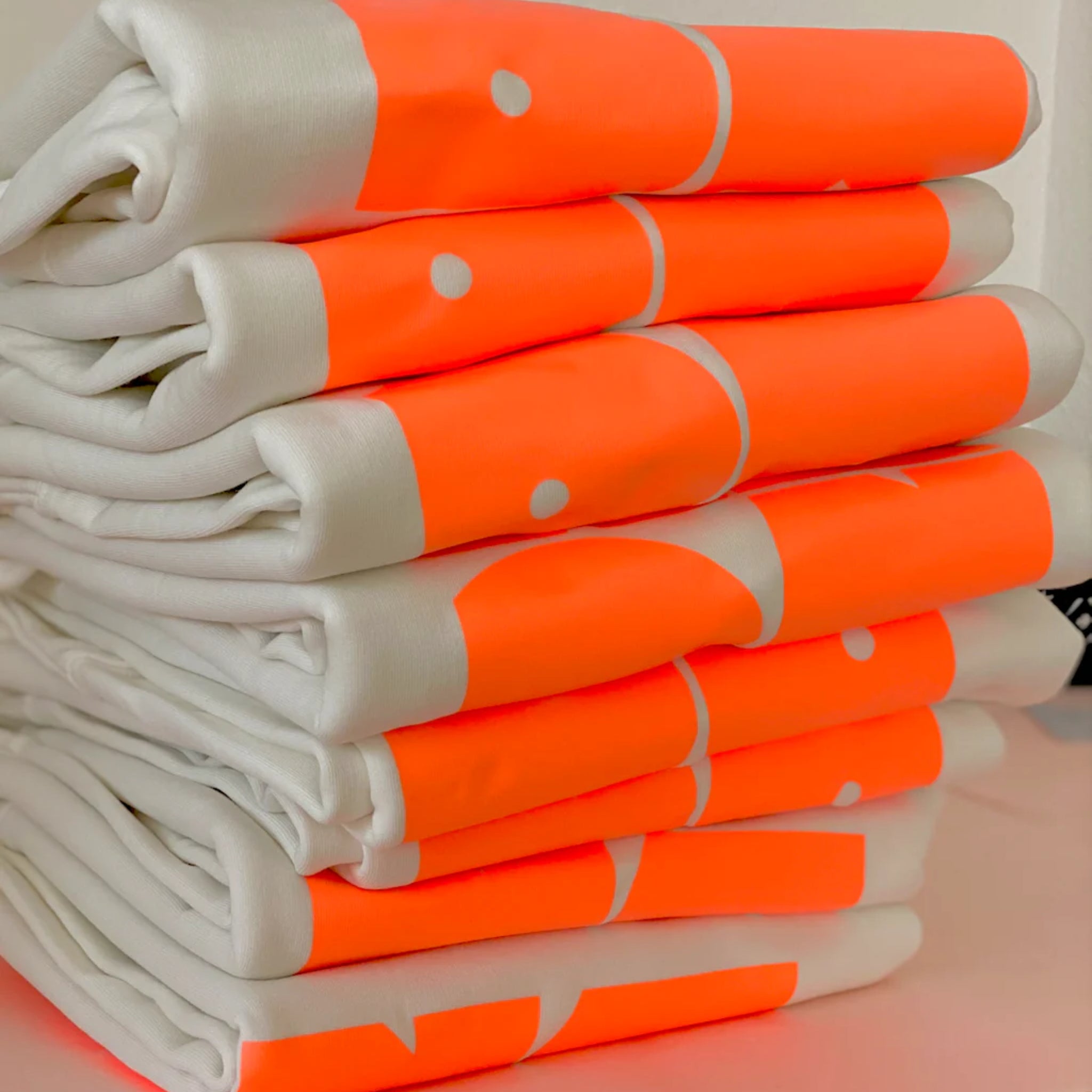 Ein ordentlich gestapelter Stapel weißer T-Shirts mit einem kräftigen, leuchtend orangefarbenen geometrischen Muster mit weißen Punkten und Kurven, die teilweise auf den gefalteten Hemden zu sehen sind.