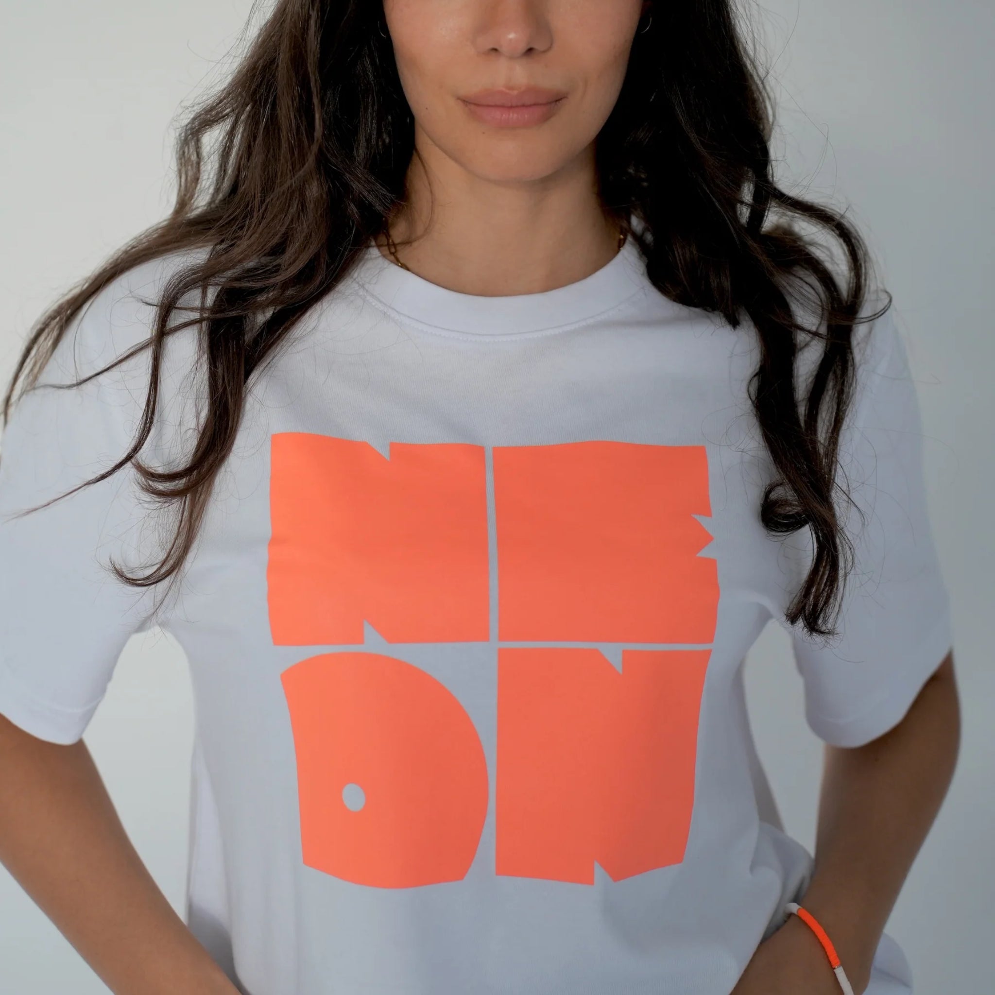 Frau mit langen dunklen Haaren trägt ein weißes T-Shirt mit großen, fetten, leuchtend orangefarbenen Buchstaben, auf denen "NEON" in einer Blockschrift steht. Das Bild ist an den Schultern und der Körpermitte abgeschnitten und hat einen neutralen Hintergrund.