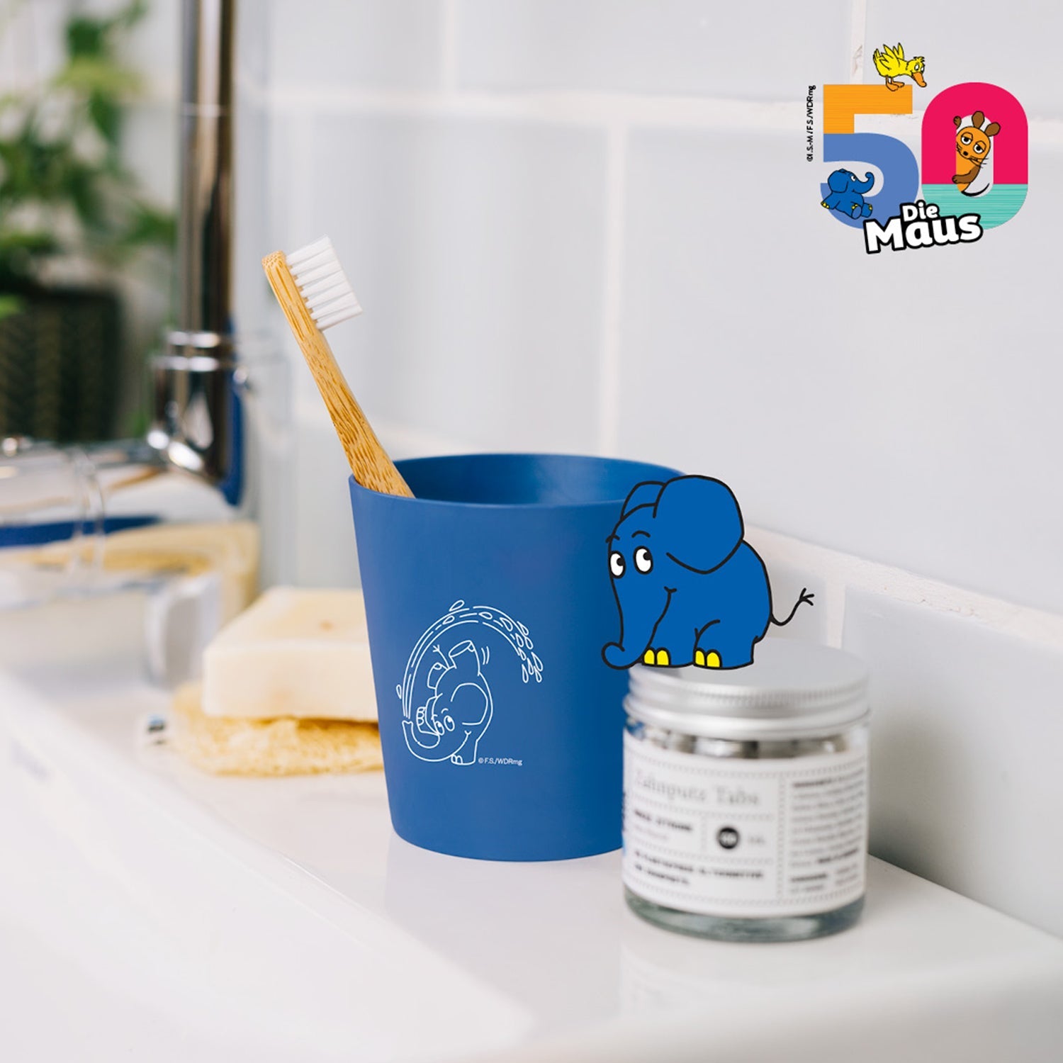 Blauer Zahnputzbecher mit Holzzahnbürste auf einem Waschbecken, daneben Zahnputztabletten und ein Elefant-Illustration.