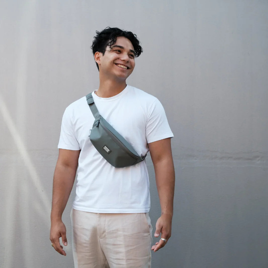 Ein lächelnder Mann mit lockigem Haar, der ein weißes T-Shirt, eine helle Hose und eine graue Umhängetasche trägt, steht vor einer schlichten grauen Wand.