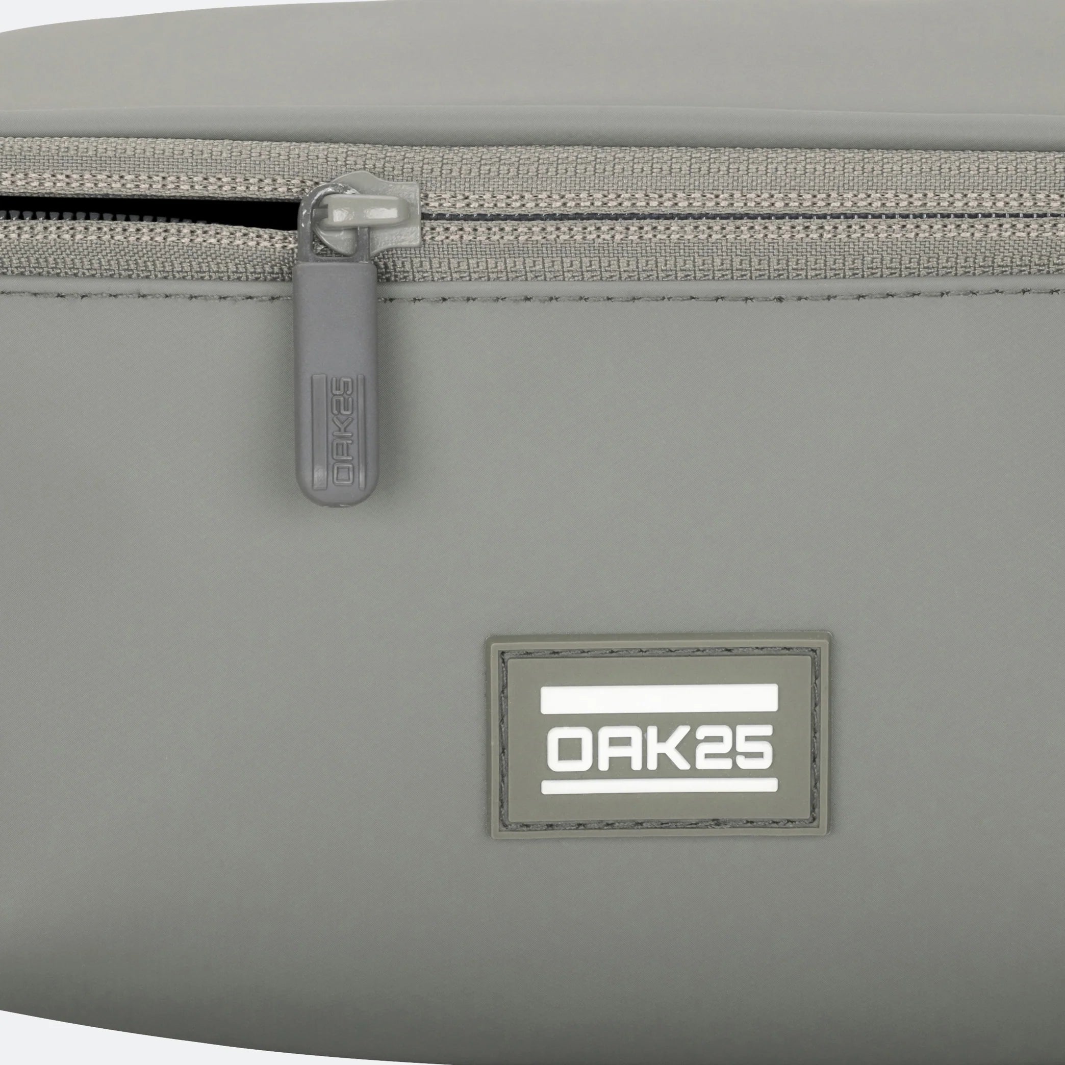 Nahaufnahme einer grauen Tasche, die einen Reißverschluss und ein rechteckiges Etikett mit dem Text OAK25 in weißen Buchstaben auf einem reflektierenden Hintergrund zeigt.