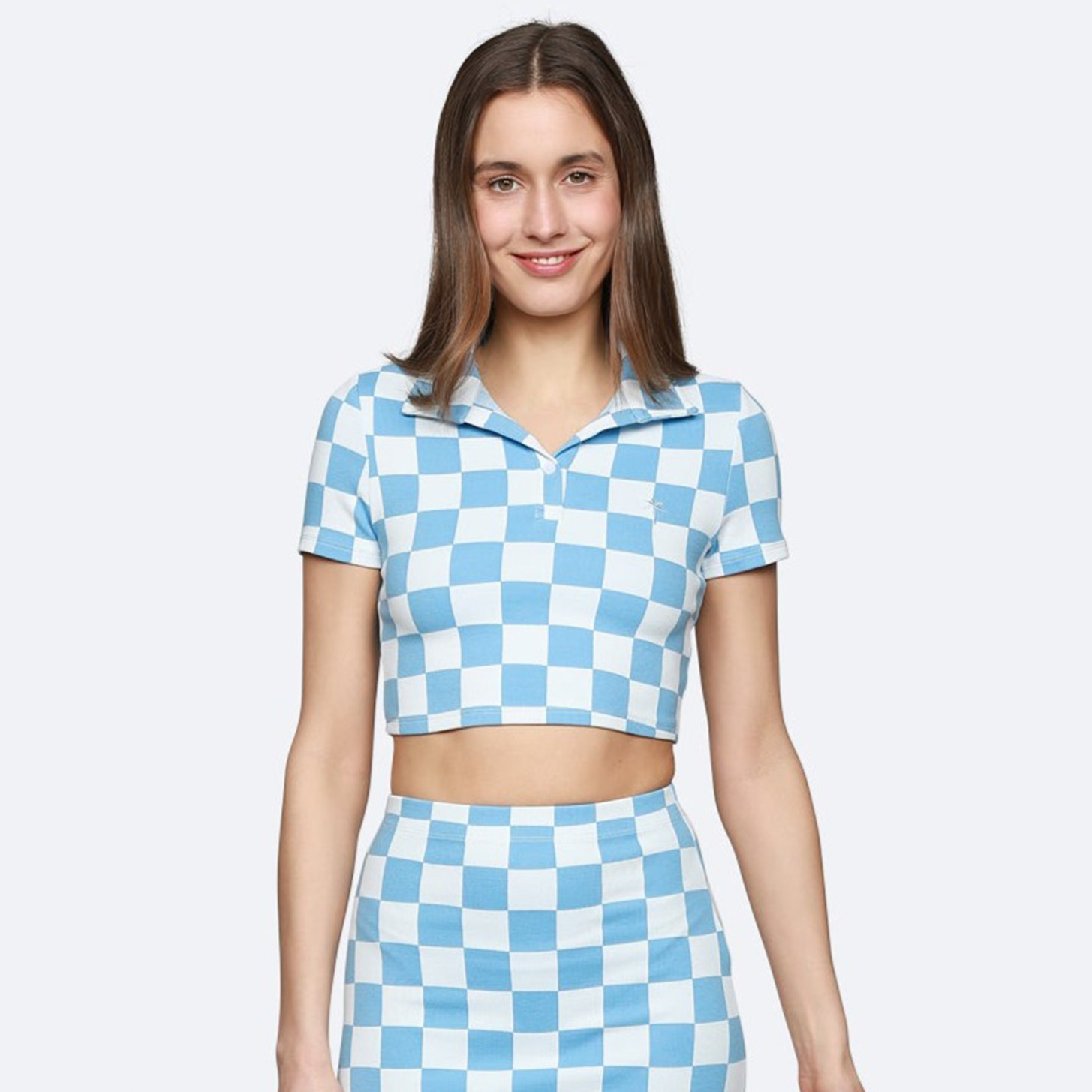 Eine Frau steht lächelnd da und trägt ein passendes hellblau-weiß kariertes Crop-Top mit Kragen und einen hochtaillierten Minirock. Sie hat glattes braunes Haar und ist vor einem schlichten hellen Hintergrund abgebildet.
