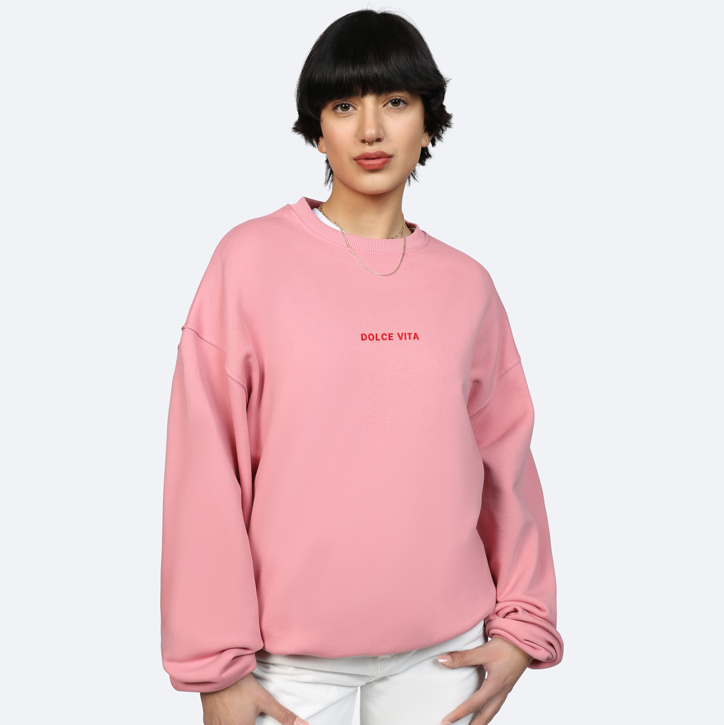 Eine Person mit kurzen dunklen Haaren trägt ein rosafarbenes Sweatshirt mit der Aufschrift DOLCE VITA und eine weiße Hose. Sie posiert mit in die Hüften gestützten Händen vor einem schlichten hellen Hintergrund.
