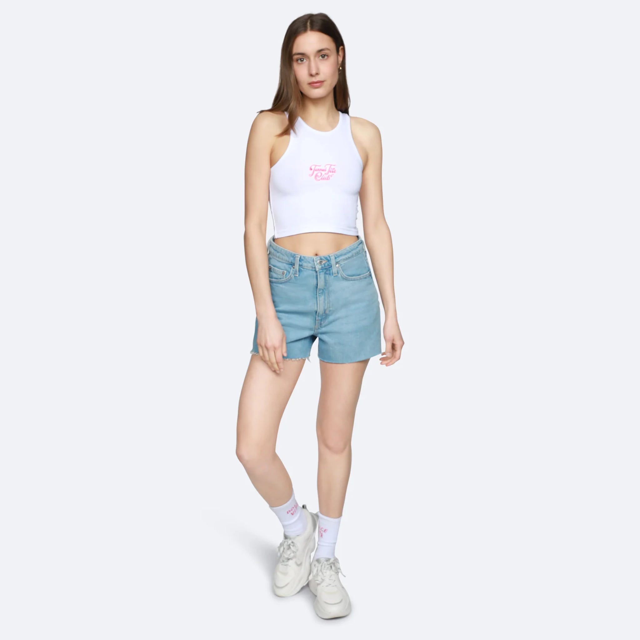 Frau in einem weißen Tanktop und blauen Shorts steht vor einem grauen Hintergrund.