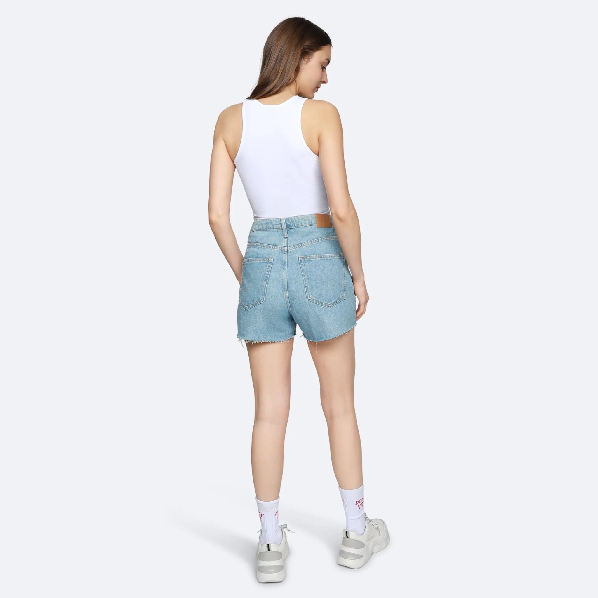 Frau im weißen Tanktop und blauen Jeansshorts, die sich von hinten zeigt, vor grauem Hintergrund.